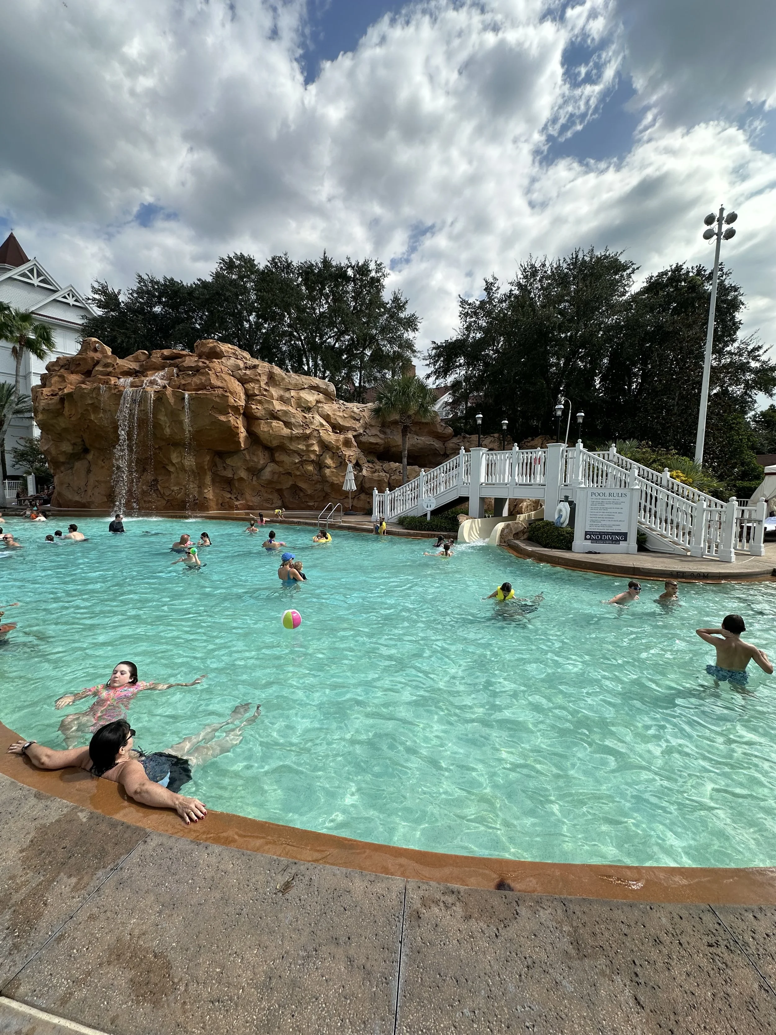 People swimming and relaxing in a large outdoor pool with a rock waterfall feature and a white bridge.