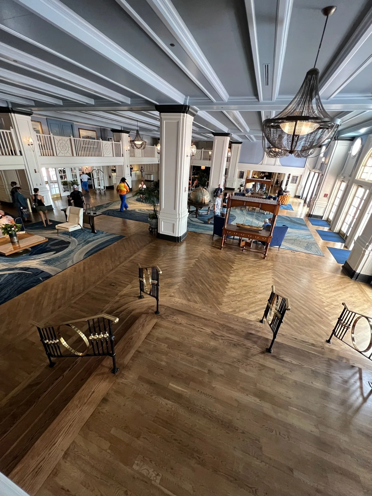 Hotel lobby with a high ceiling, large chandelier, polished wooden floors, and large windows. There are a few chairs, tables, rugs, and people walking or sitting around.