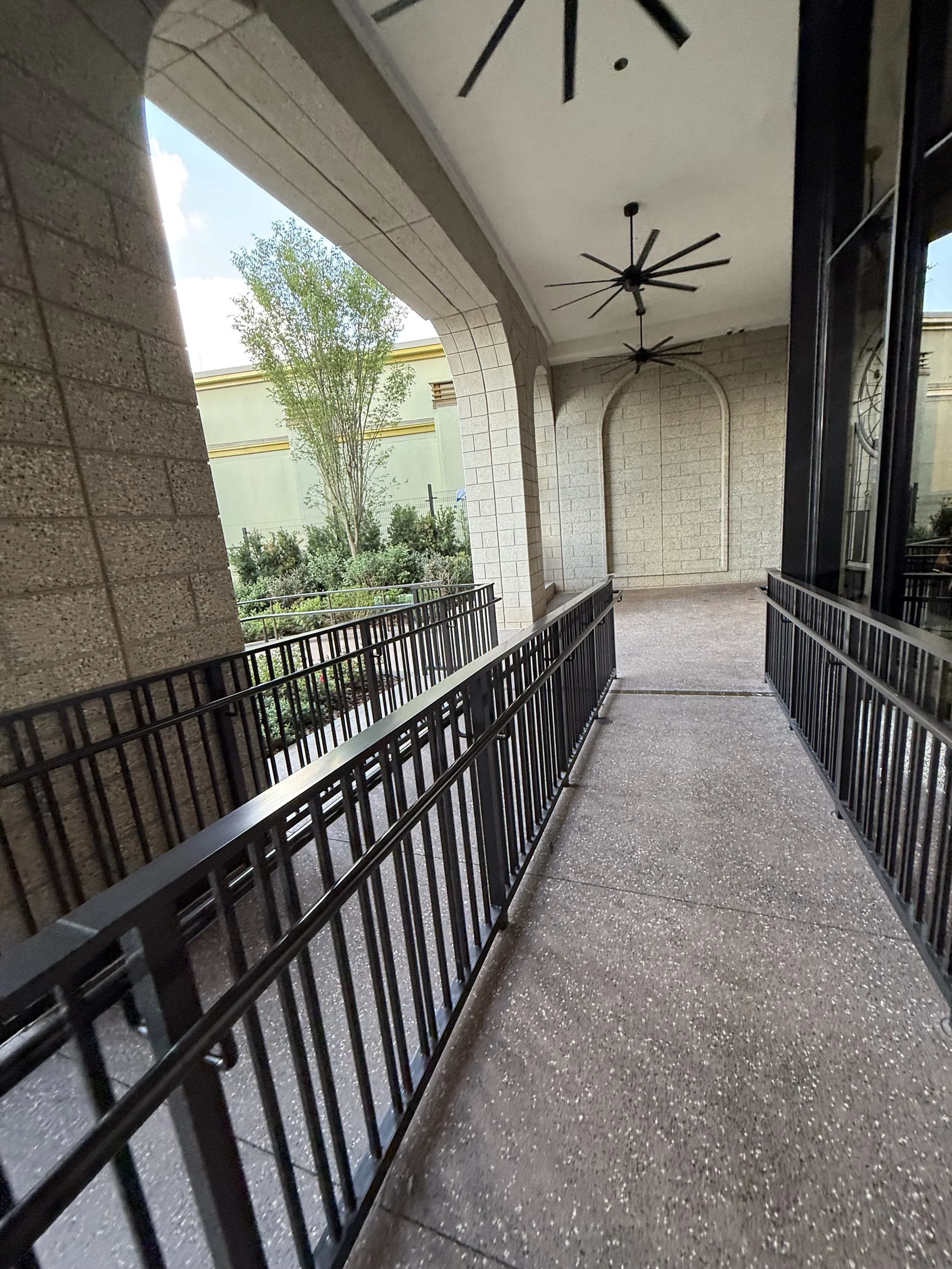 Outdoor covered walkway with black metal railings, brick walls, ceiling fans, and a decorative arch, with green trees and a building in the background.