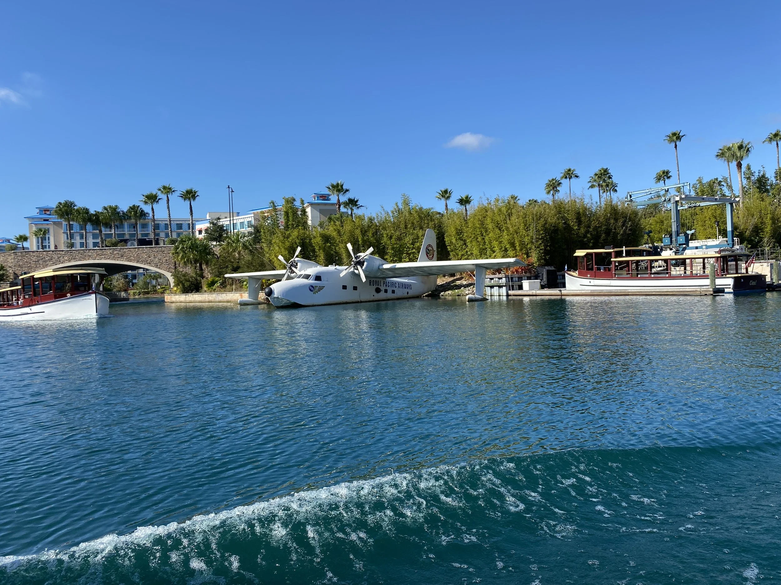 A waterway with three boats docked along the shore, including a white seaplane labeled 'Royal Pacific Airways,' and palm trees and buildings in the background under a clear blue sky.