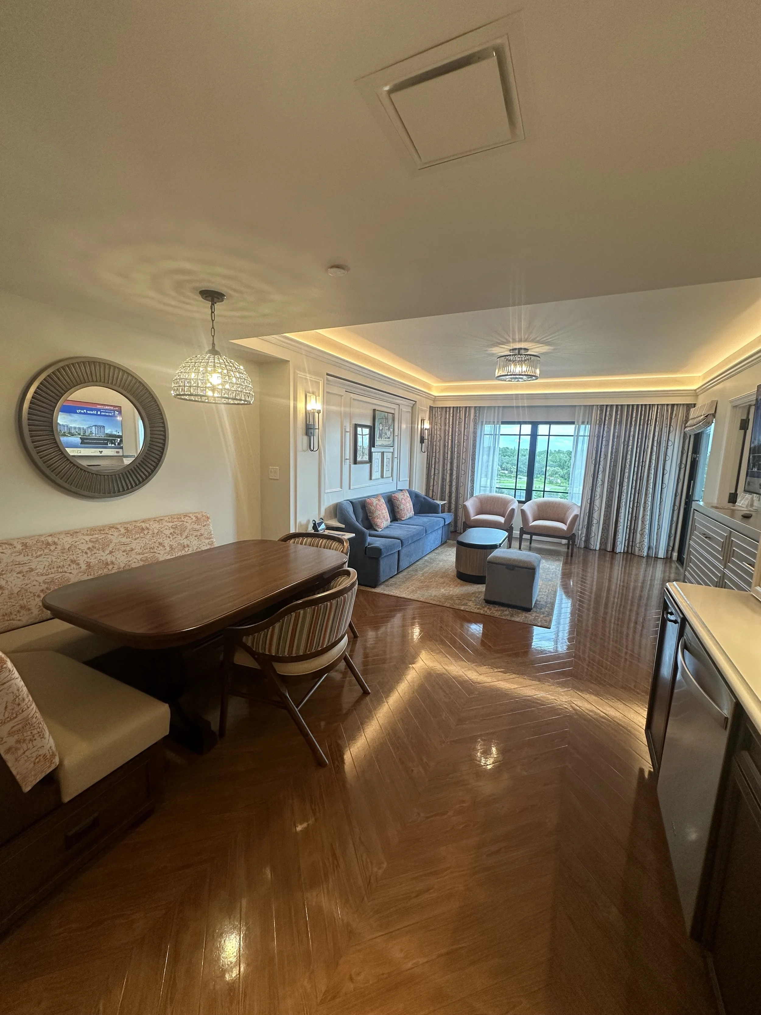 Living room with blue sofa, beige and pink armchairs, wooden dining table, patterned curtains, parquet flooring, and a window with a view outside.
