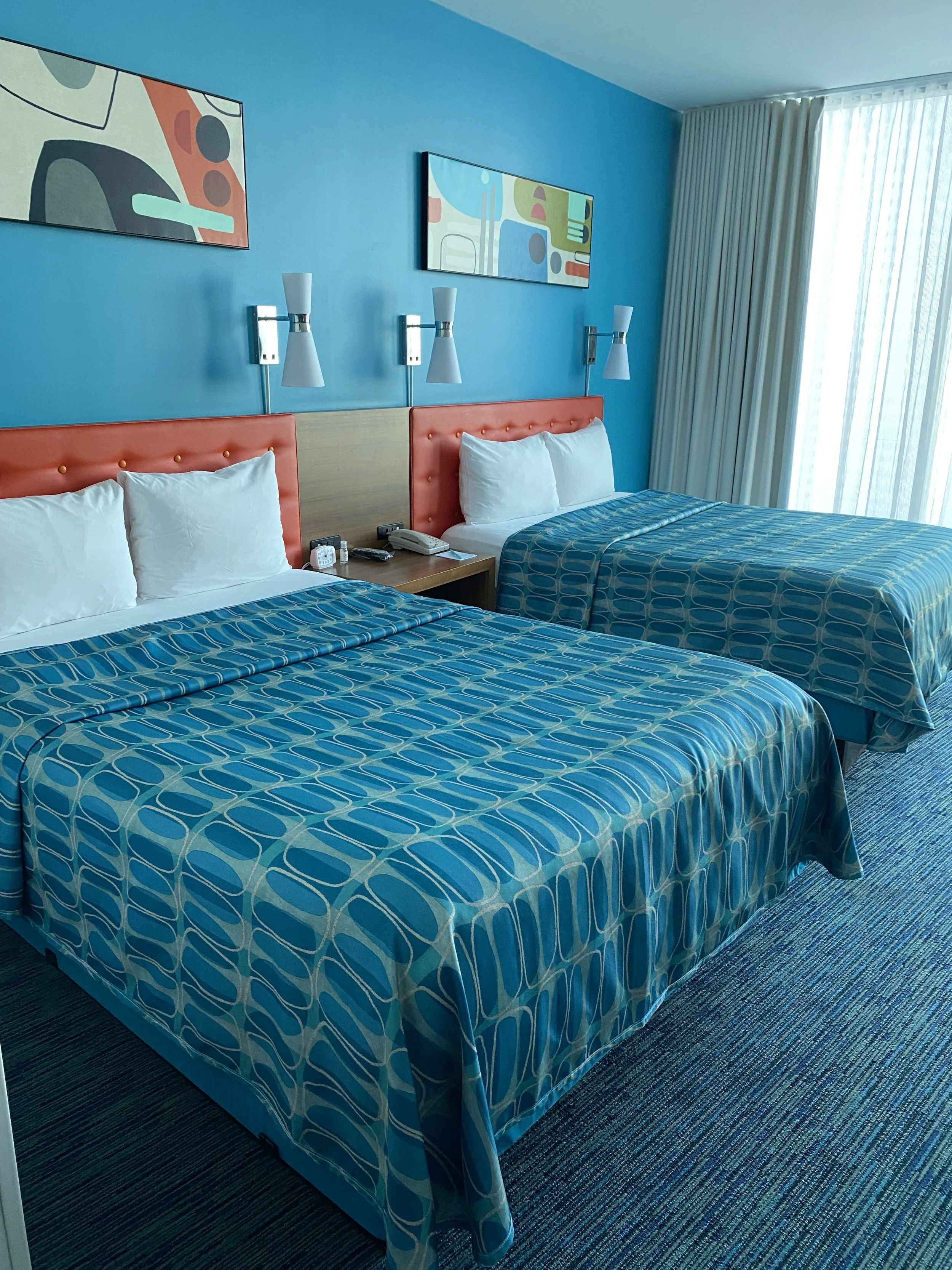 Two double beds with colorful patterned bedspreads, orange headboards, and white pillows in a hotel room. A nightstand with a telephone, alarm clock, and other items is between the beds. The wall has two abstract artworks and four wall-mounted lights