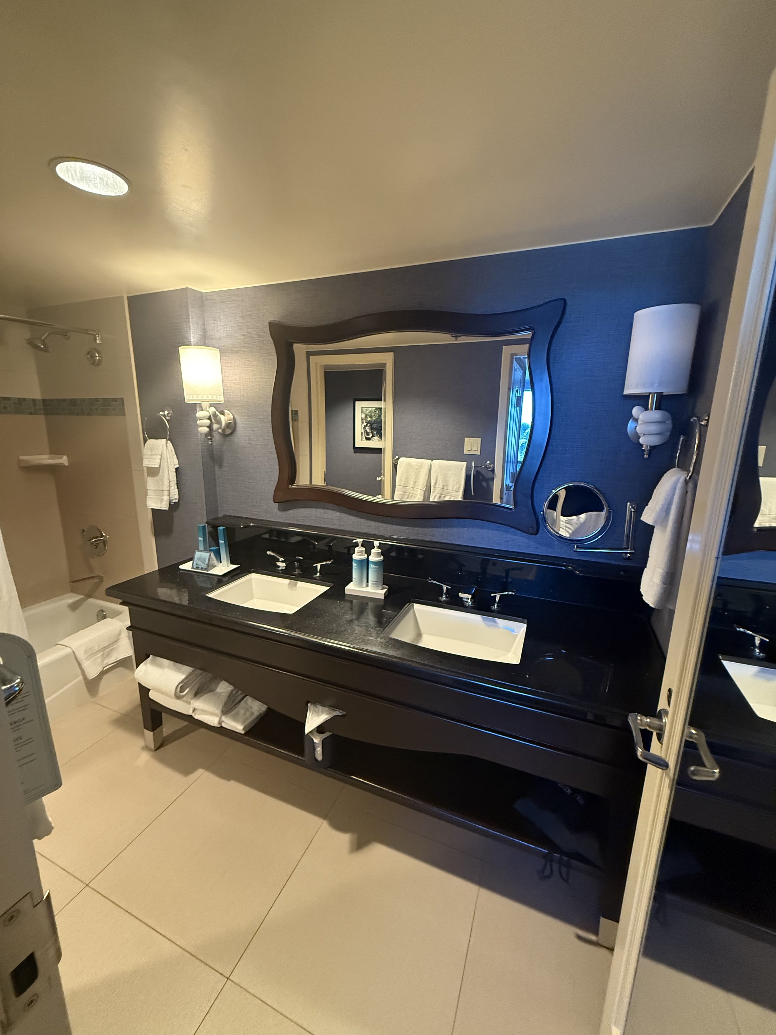 Modern bathroom with double sink vanity, black countertop, large mirror, wall-mounted lamps, towels, and a bathtub with shower behind a glass door.