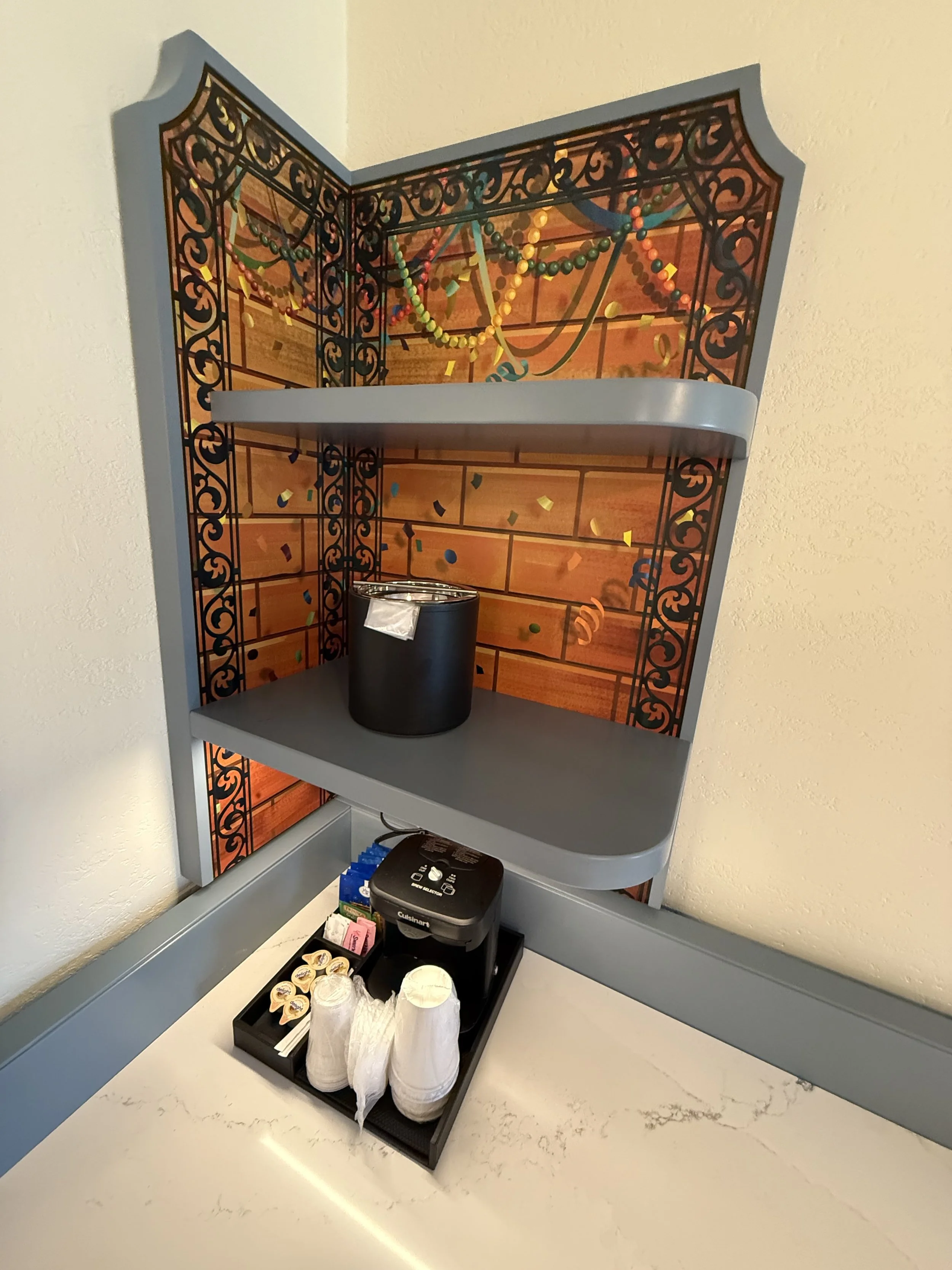 A beverage station with a black coffee machine, disposable cups, and condiments on a tray. Behind is a decorative wall panel with a brick background and Mardi Gras beads and confetti.