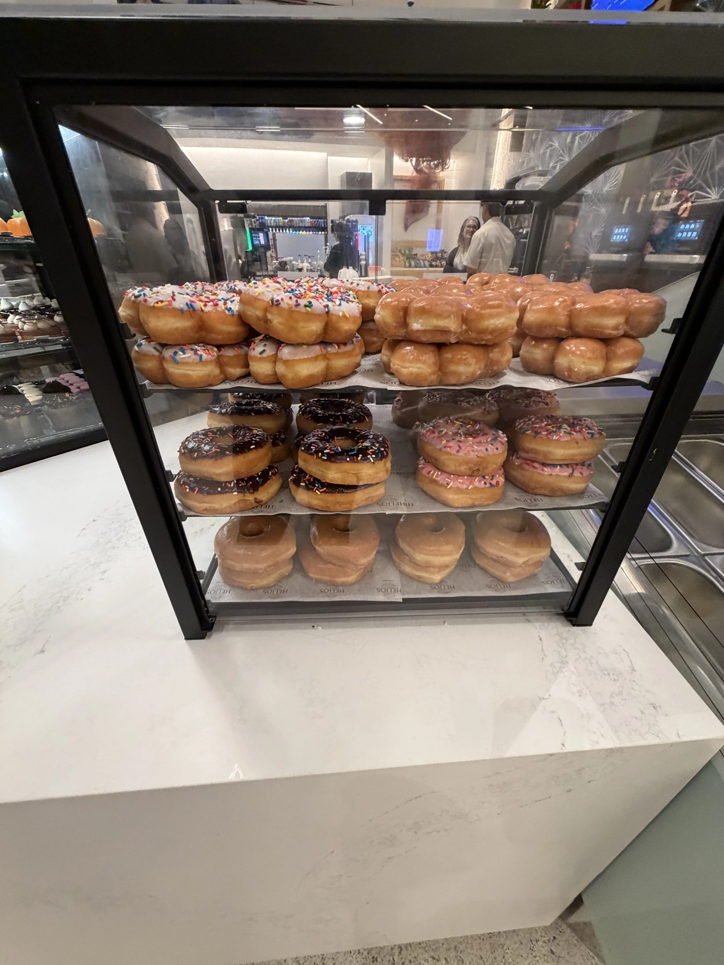 A display case filled with assorted donuts, including sprinkled, chocolate-topped, and glazed donuts, on a white countertop in a bakery or cafe.