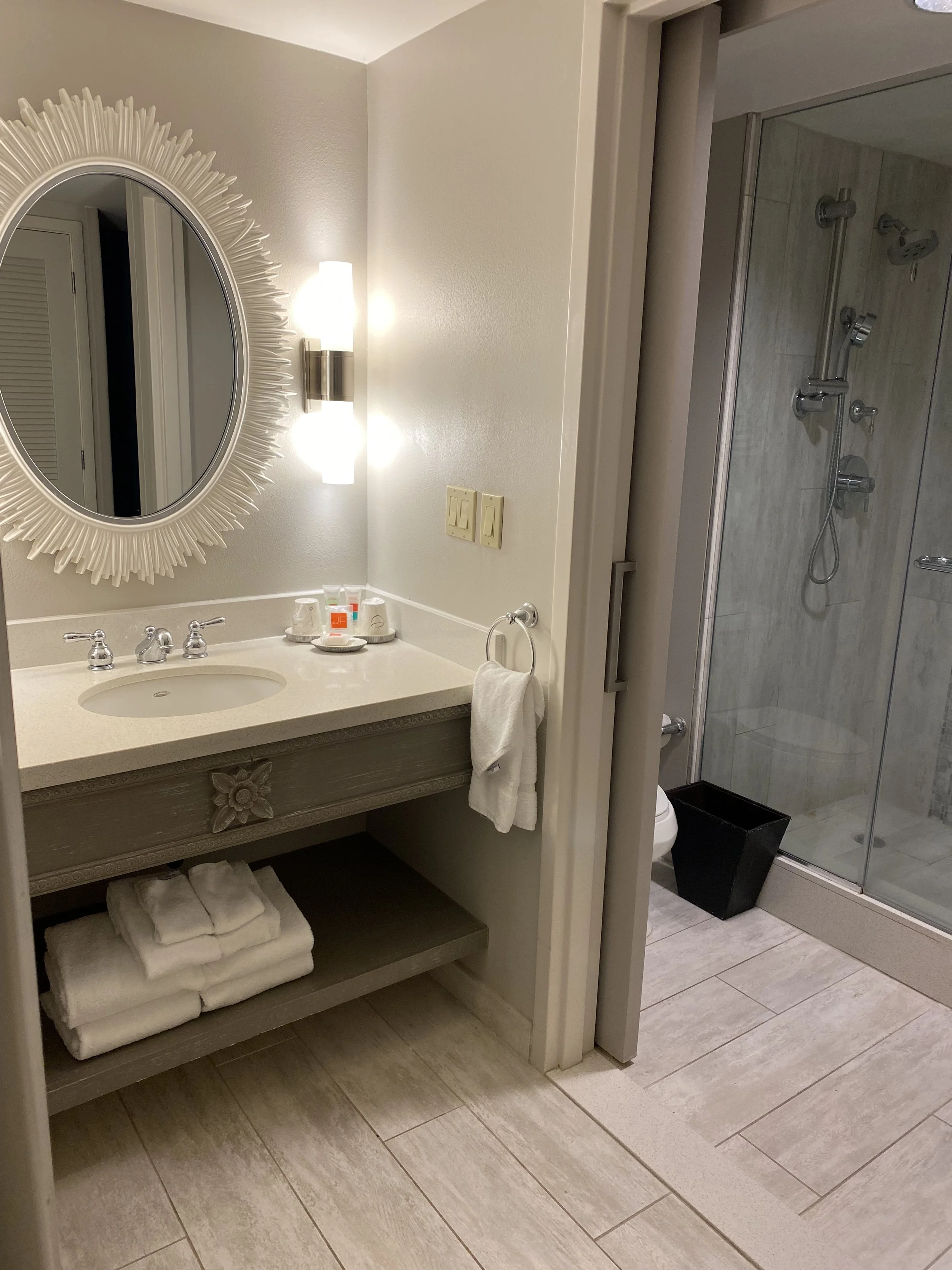 Bathroom with a decorative round mirror, a wall-mounted light, a sink with toiletries, a towel ring with a white towel, a shelf with folded towels, a shower with glass door, a black trash bin, and tiled floor.