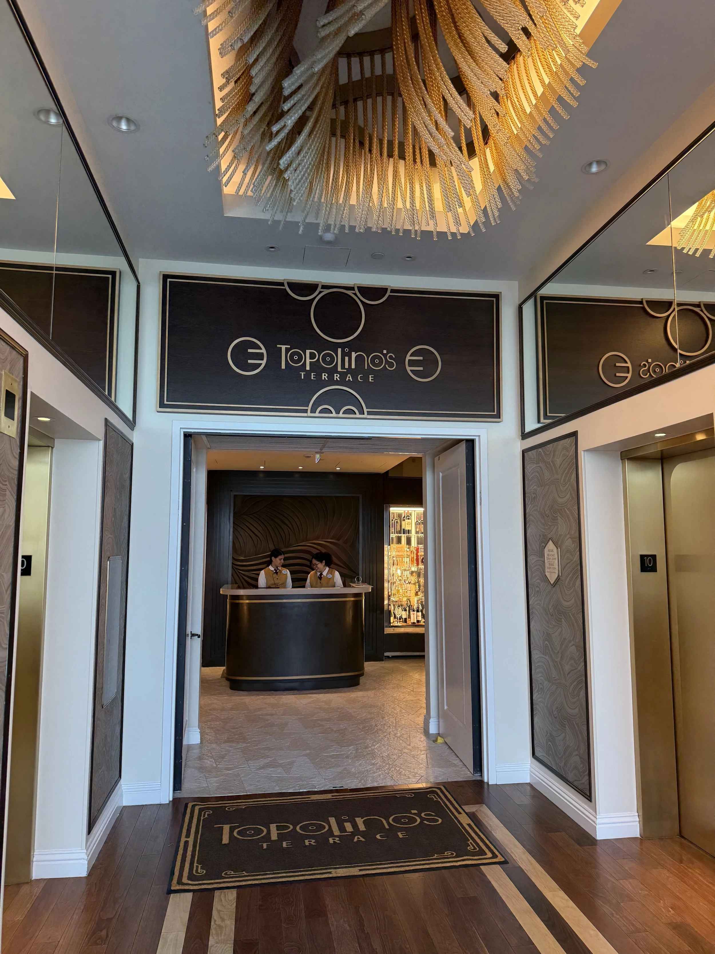 The entrance to Topolino's Terrace, featuring a black and gold reception desk with two staff members behind it. The decor includes brown, gold, and black accents, with a chandelier hanging from the ceiling and mirror panels on the walls.