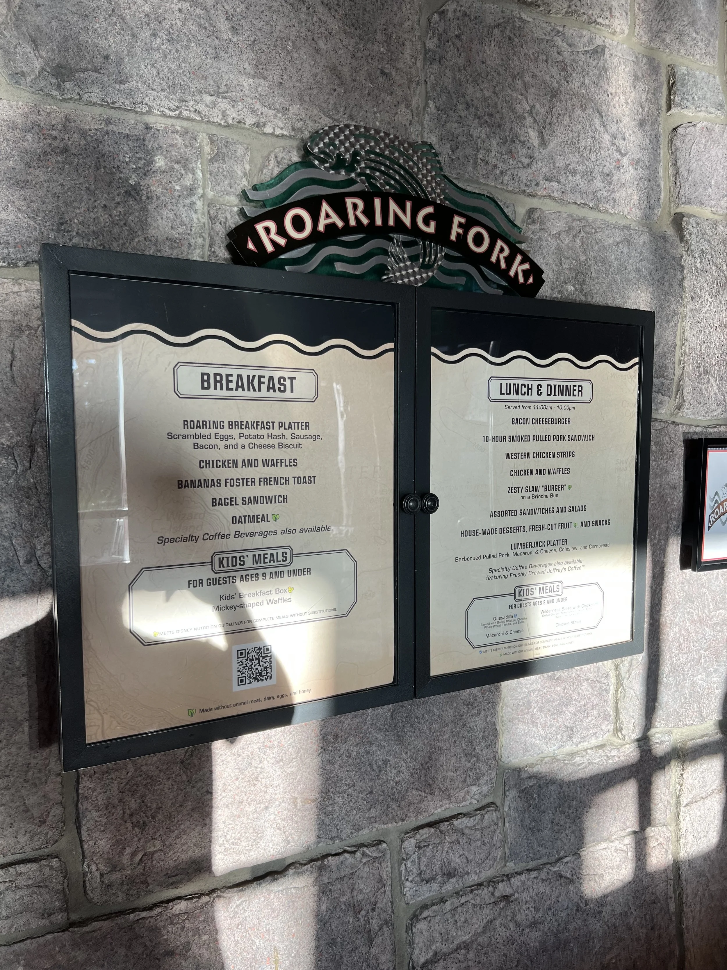 Menu board for Roaring Fork restaurant with breakfast options on the left and lunch & dinner options on the right, mounted on a stone wall. Roaring Fork sign with a forest-themed logo is above the menu.