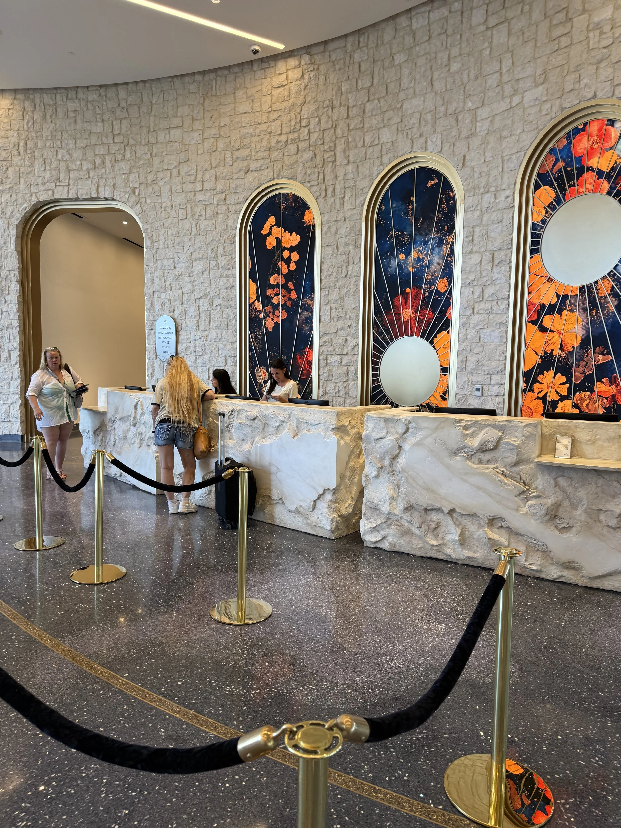 Hotel reception desk with a high stone counter and three tall arched windows featuring floral designs in orange and red. Four people are at the desk, with one woman talking to another woman and two staff members behind the counter.