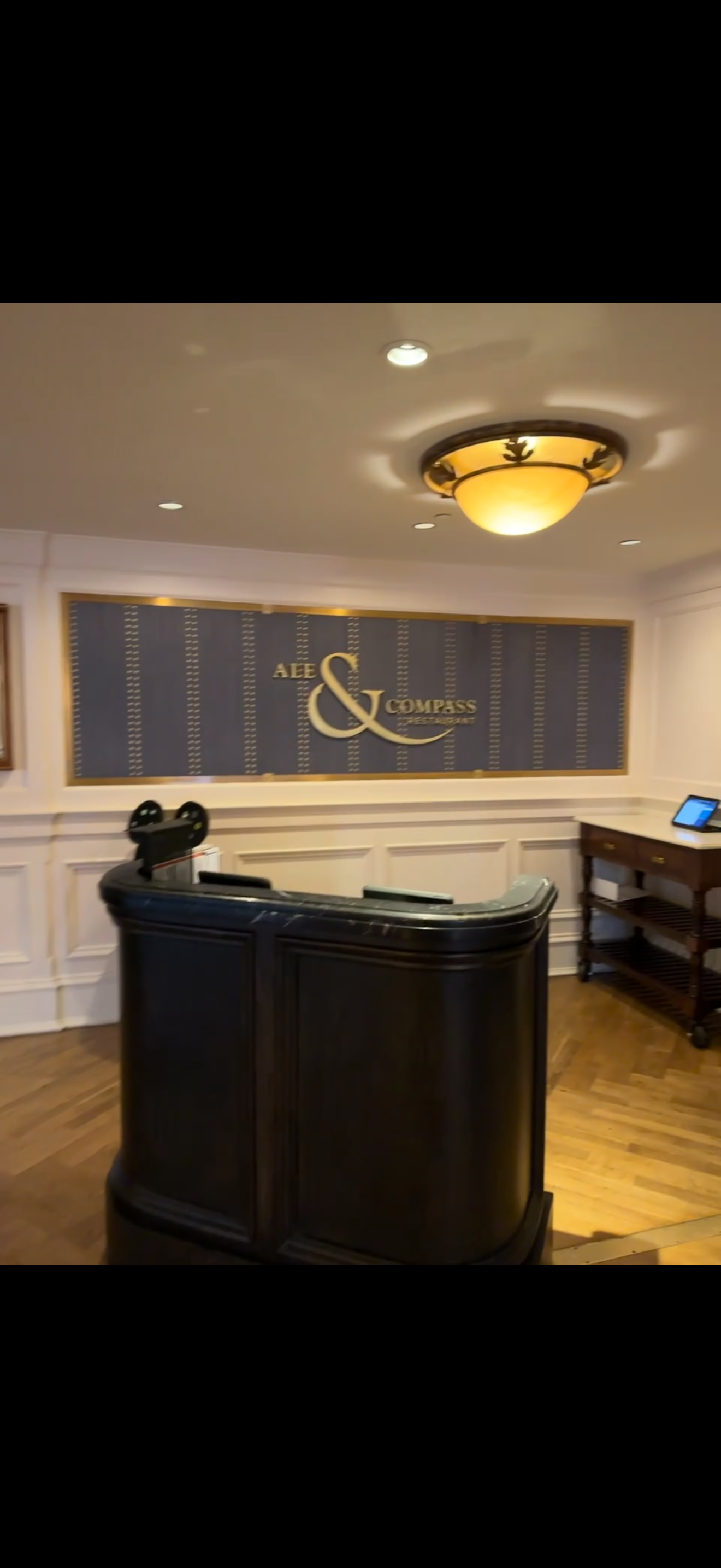 Hotel reception area with a dark curved front desk, a wall sign reading 'Alee & Compass', and a small table with a digital device on it.