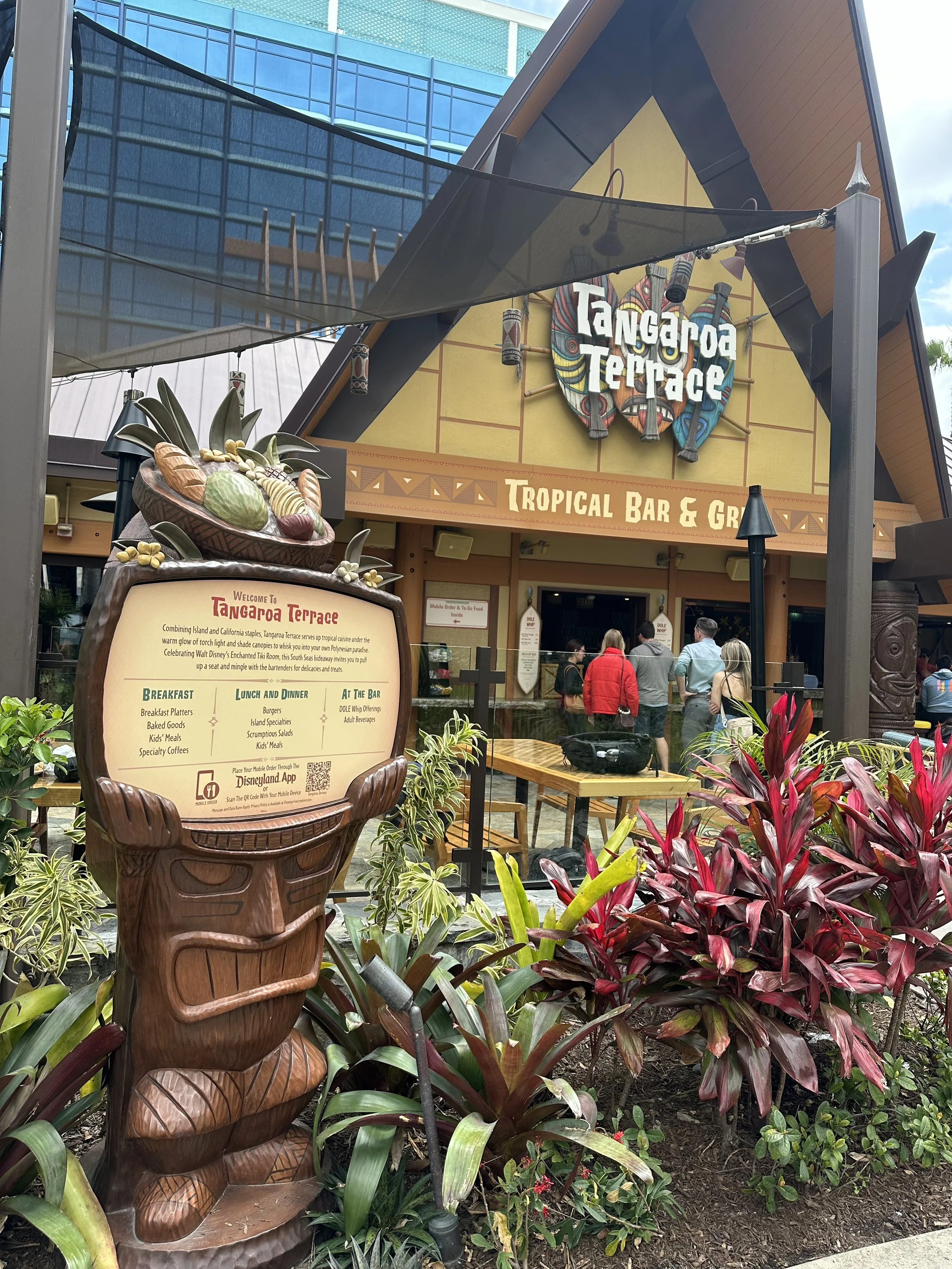 Entrance to Tangaora Terrace, a tropical bar and grill, featuring a wooden tiki-style sign with a menu and tropical plants in the foreground, with people standing in line at the entrance.