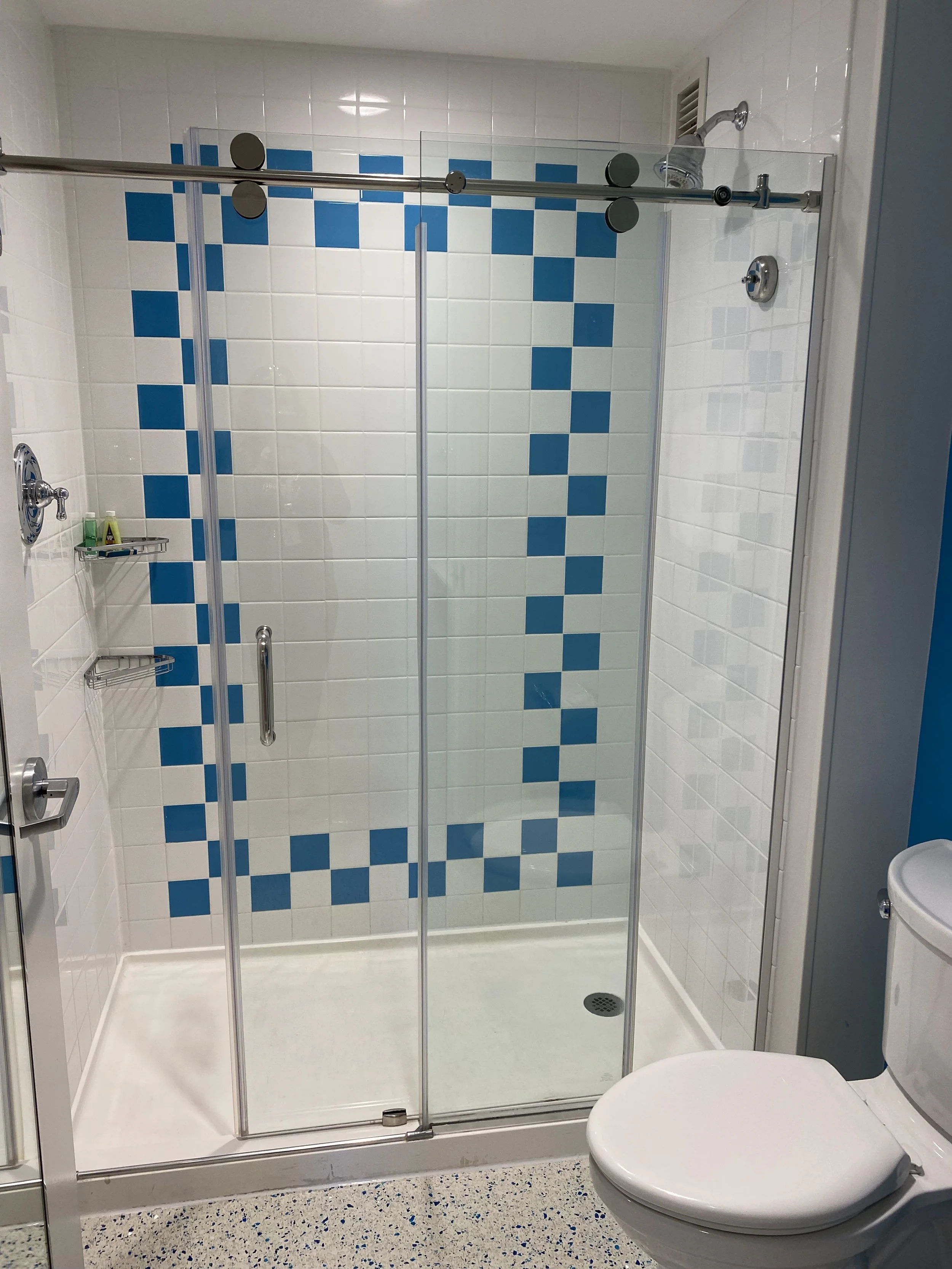 A clean shower stall with clear glass doors, blue and white tile pattern on the wall, and a showerhead, with a white toilet to the right and a textured floor.