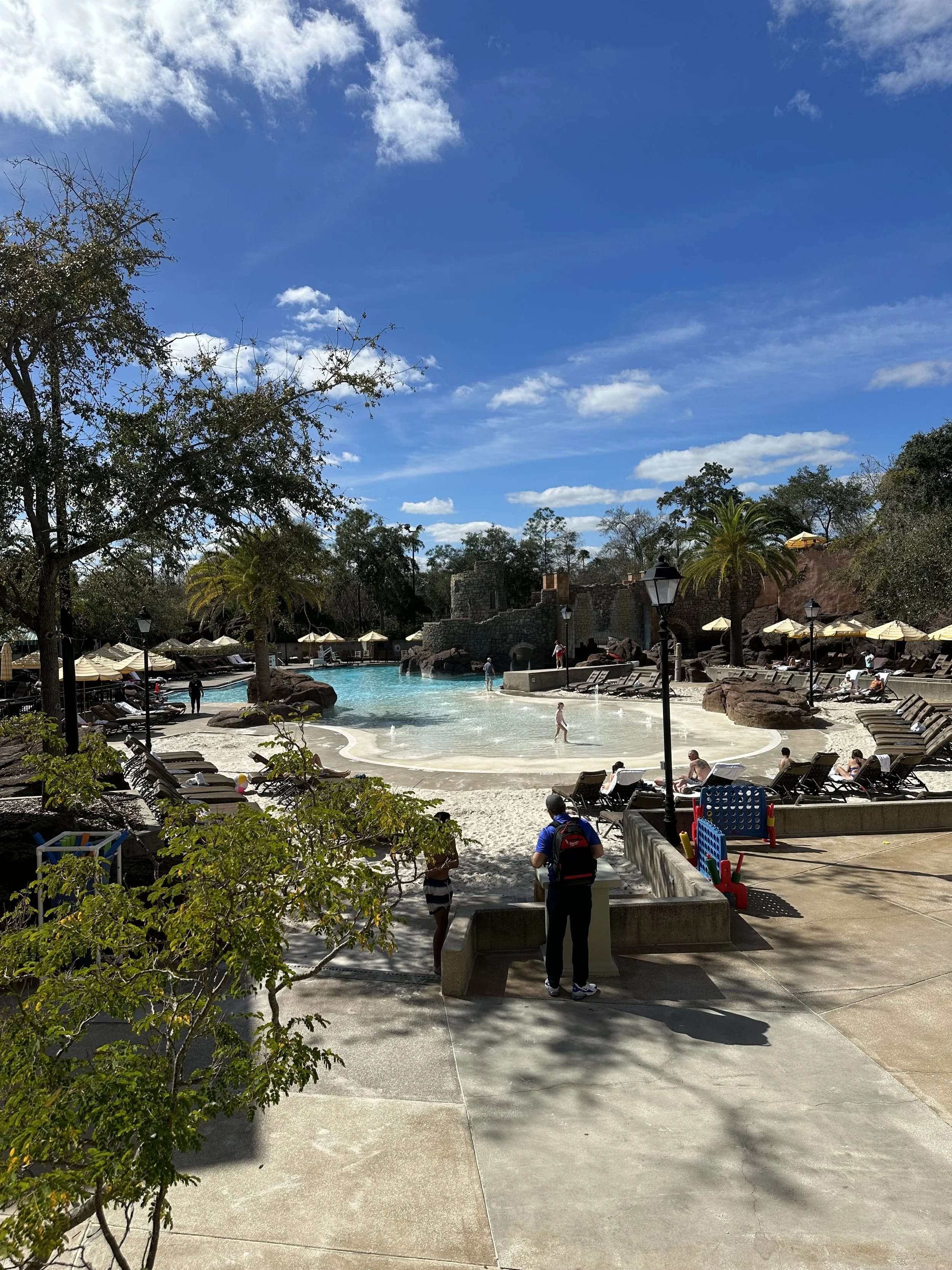 A sunny day at a resort pool with lounge chairs, umbrellas, and palm trees; people are swimming and relaxing by the pool.