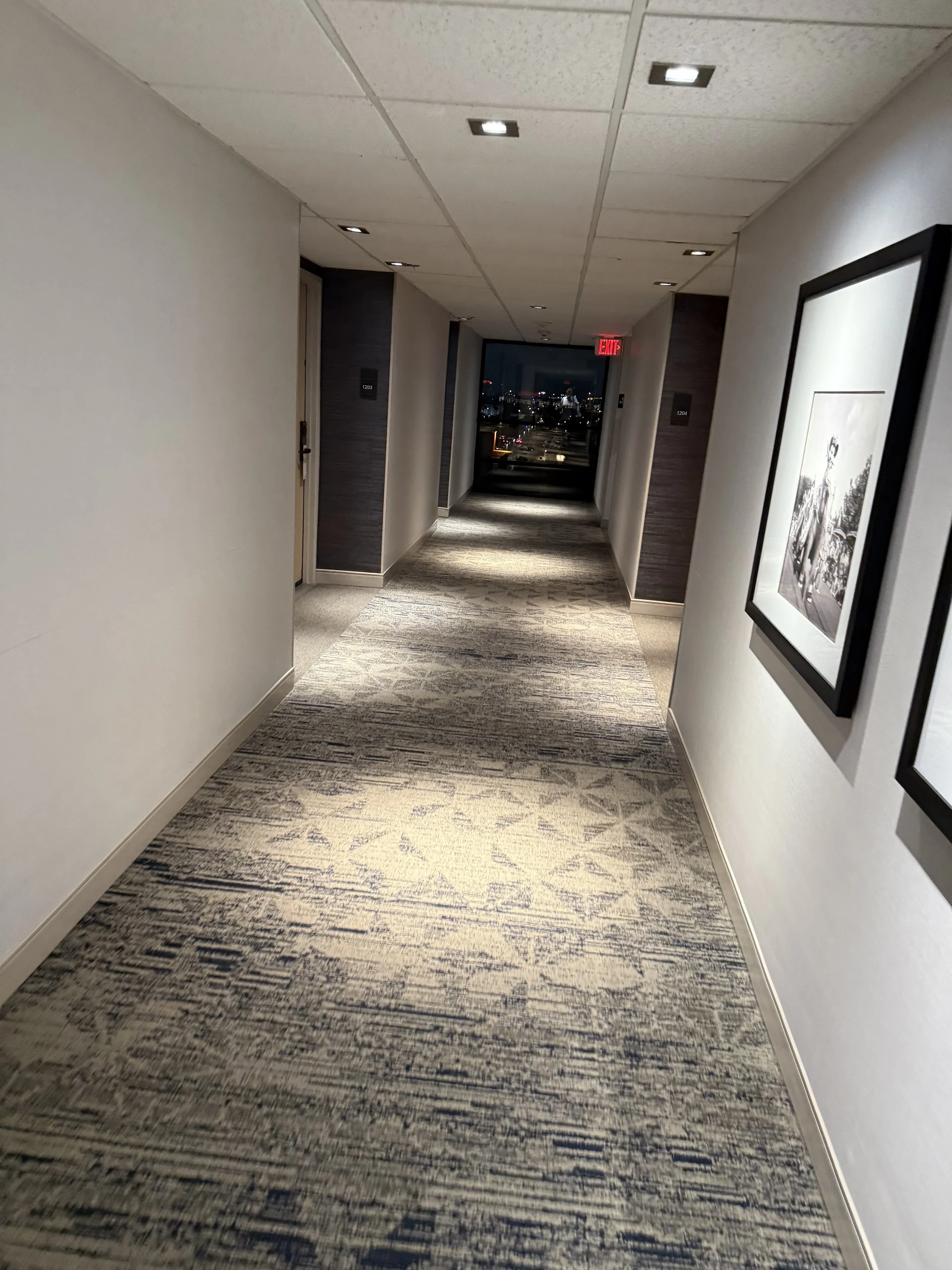 Empty hotel hallway with framed artwork on the right wall and a city view at the end of the corridor at night.