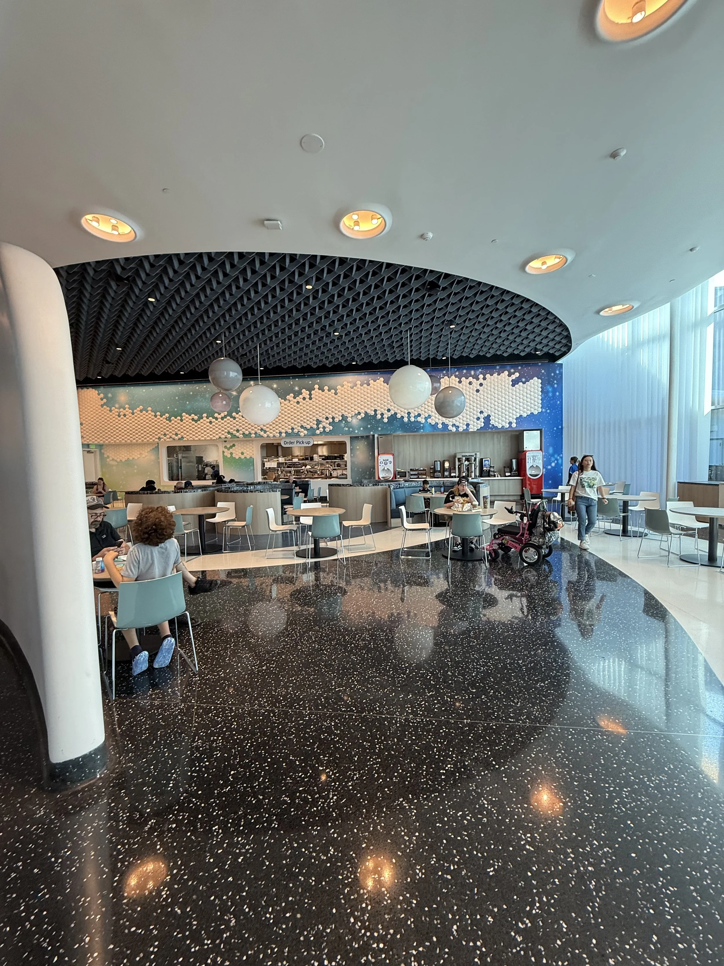 Modern airport lounge with black terrazzo floor, white and blue chairs, and a curved wall with abstract clouds painted on it. There are several people seated and standing, including a woman pushing a stroller. The ceiling has round recessed lighting 