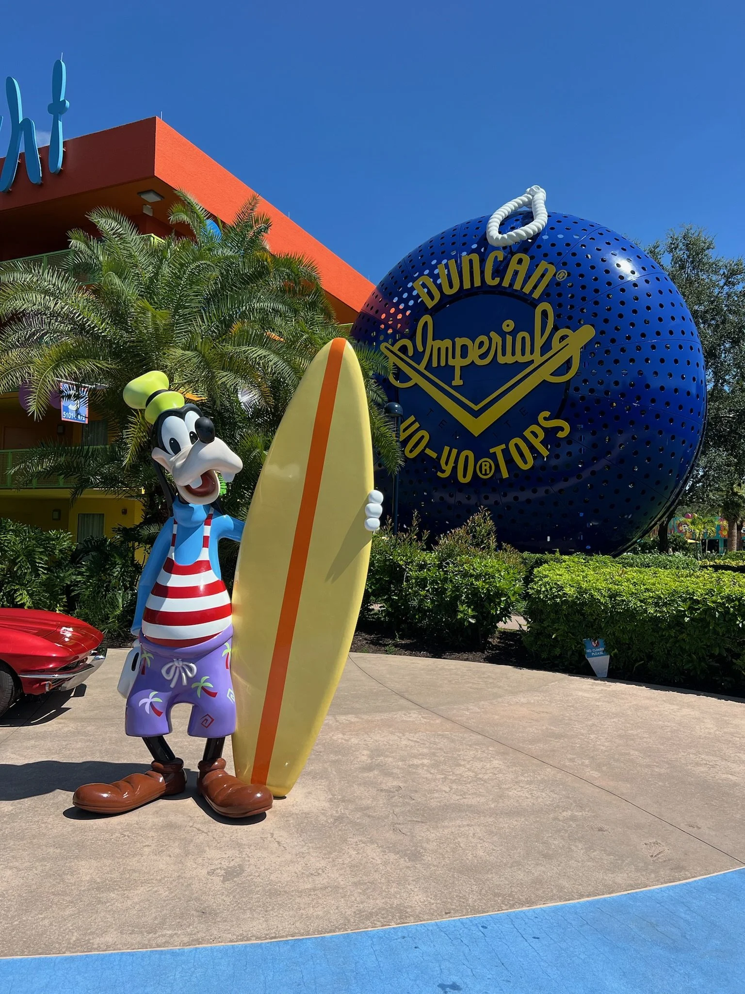 Goofy character holding a yellow surfboard outside Disney's Hollywood Studios with a replica of a golf ball and a blue sign reading "Duncan Imperial" and "You Top" in the background.