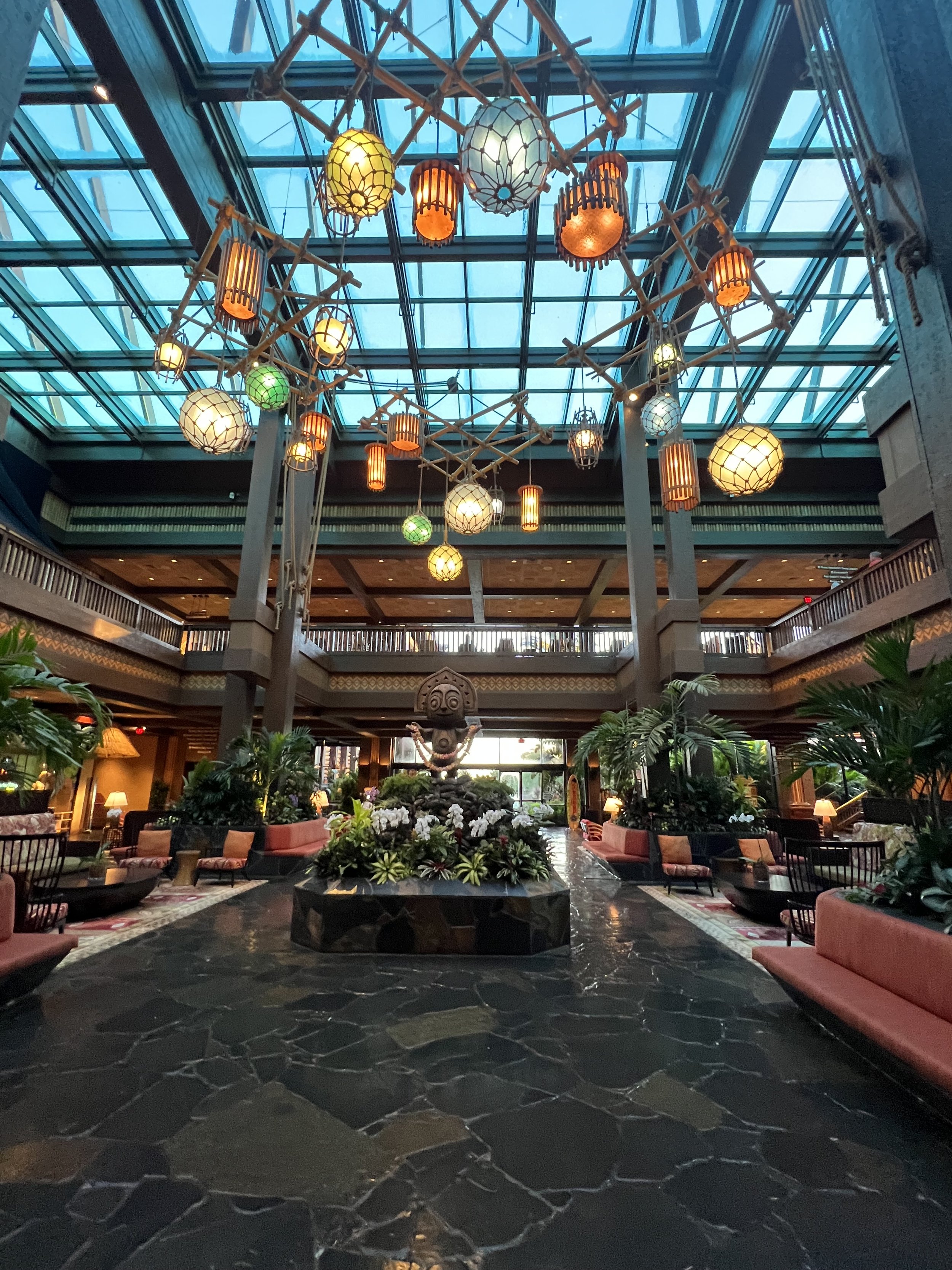 Interior of a hotel lobby with a large glass ceiling, hanging lanterns, and tropical plants surrounding a central statue.