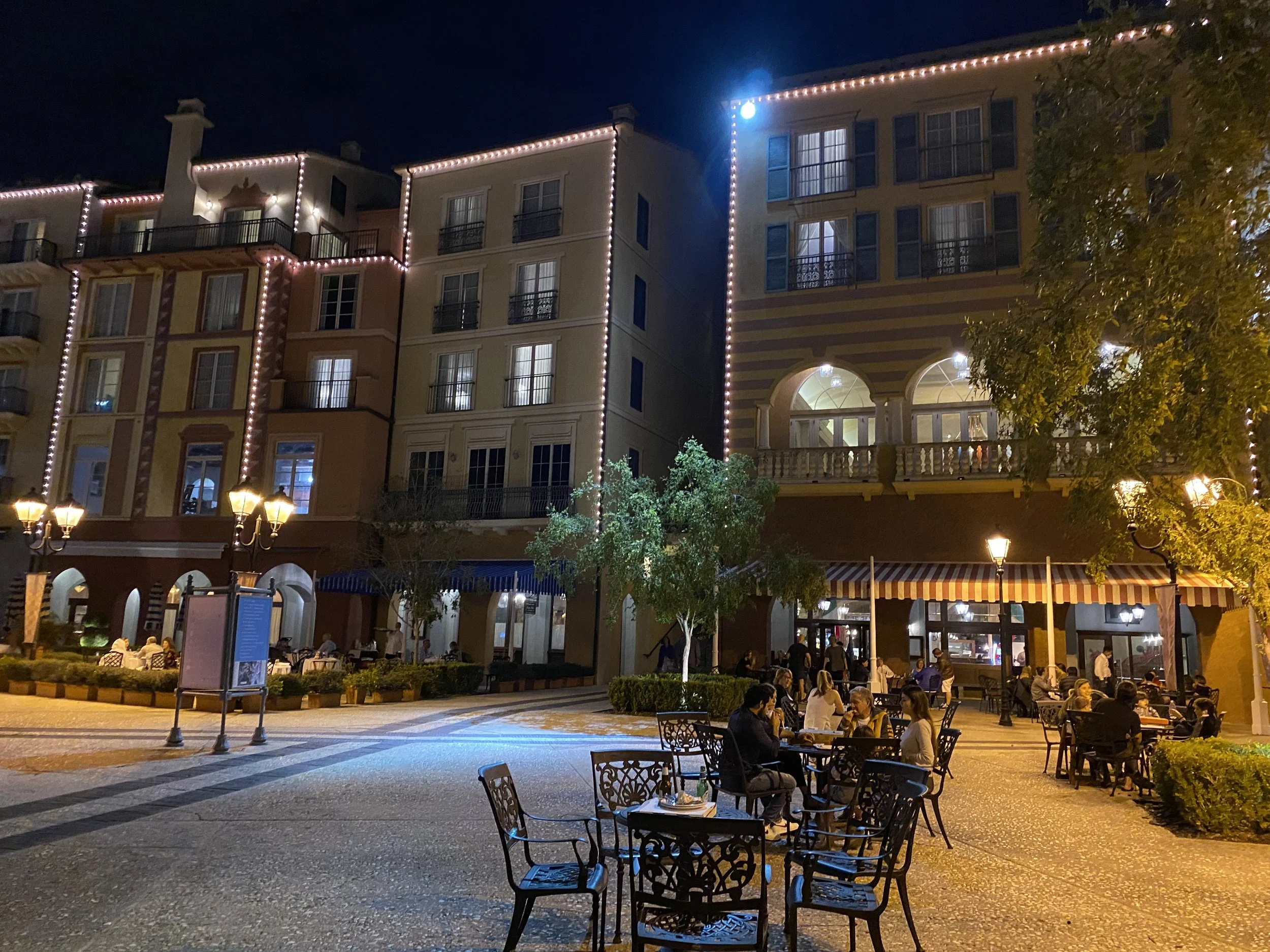 Outdoor dining area at night with tables and chairs filled with people, surrounded by multi-story buildings decorated with string lights, trees, and street lamps illuminating the area.