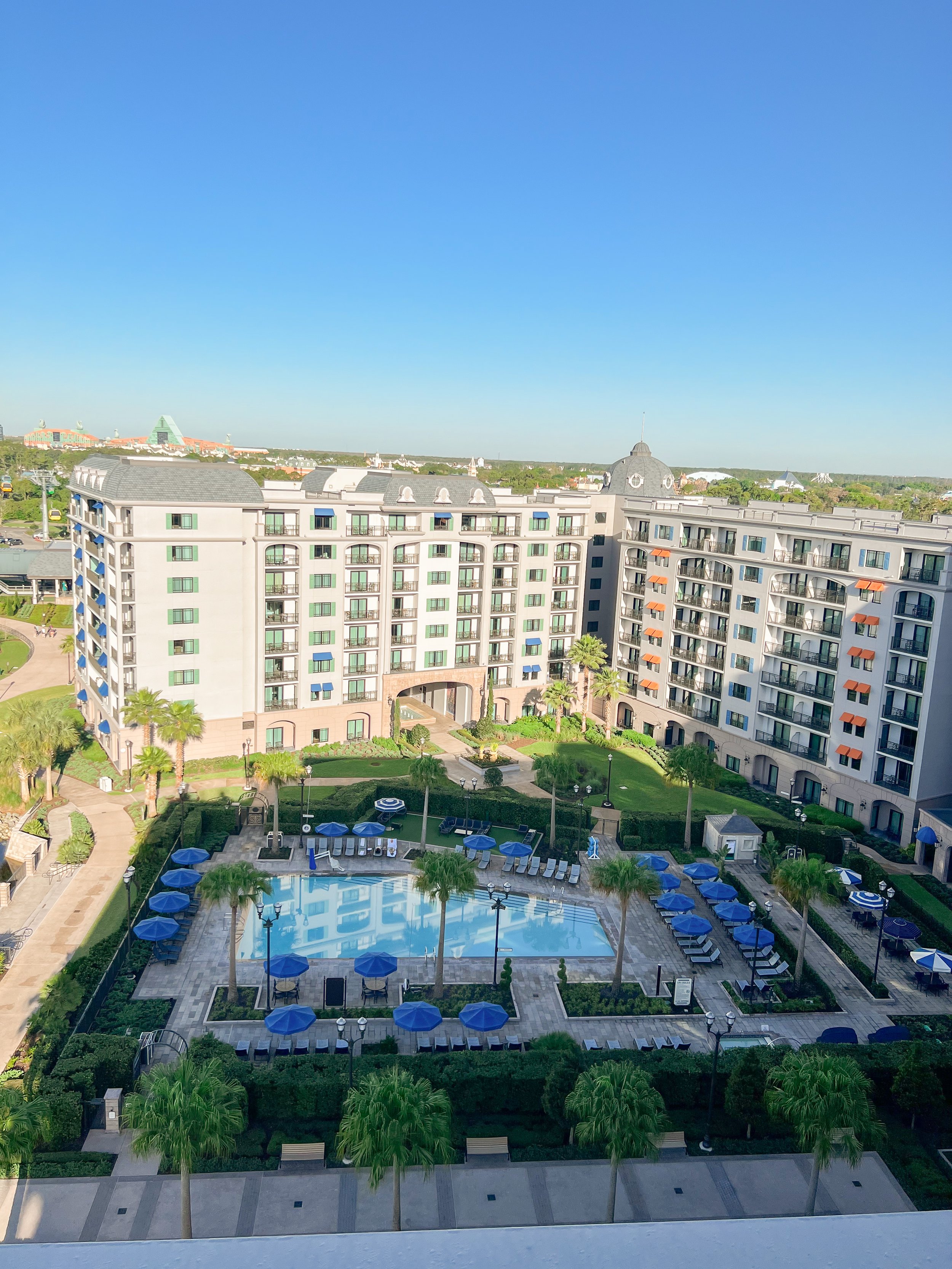 View of a residential complex with a swimming pool, surrounded by lounge chairs, umbrellas, and palm trees.