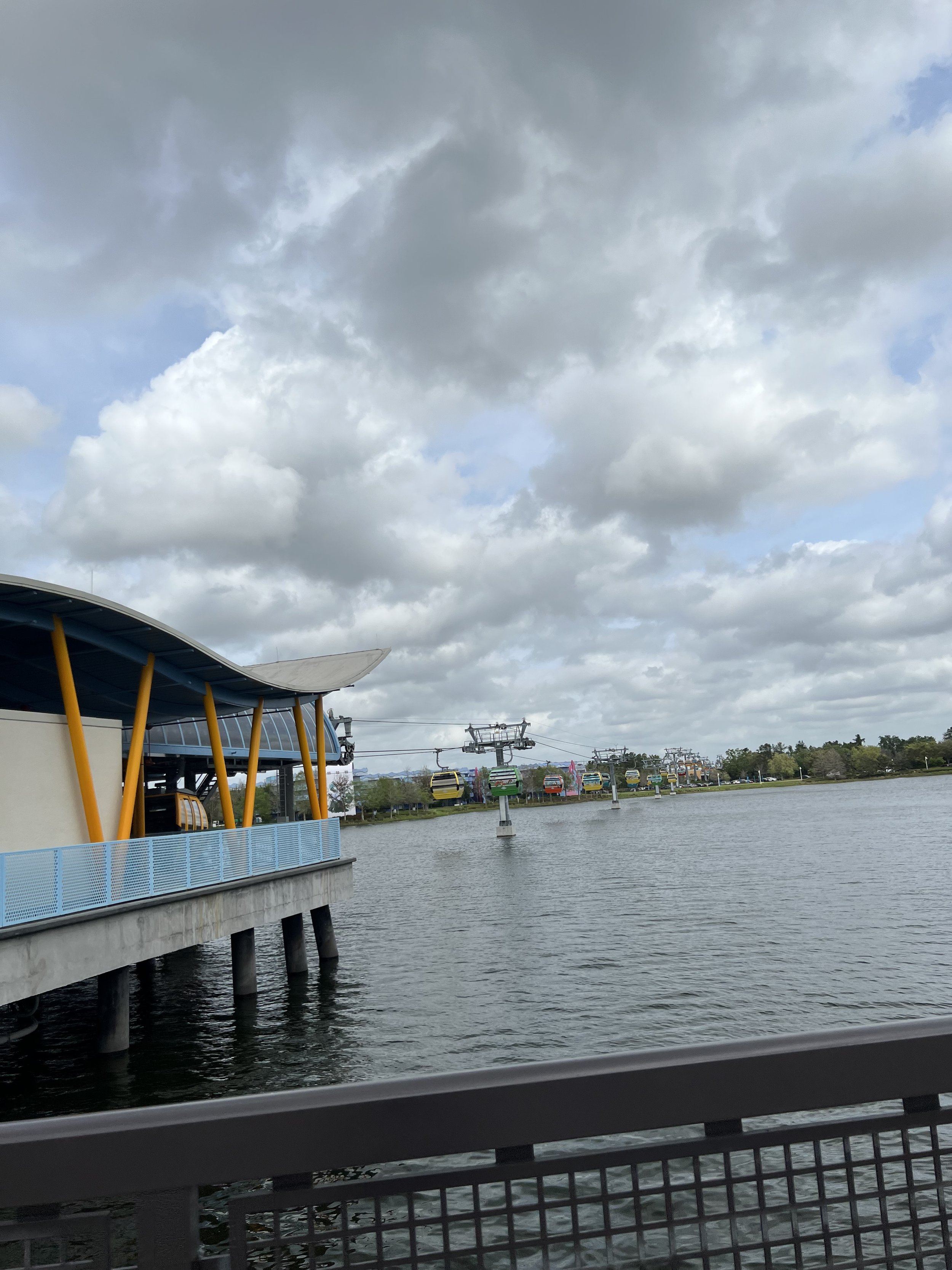 A waterfront scene with a building on the left that has yellow columns and a curved roof, a body of water in the foreground, and a series of colorful gondolas or boats in the distance on a cable car lift system under a cloudy sky.