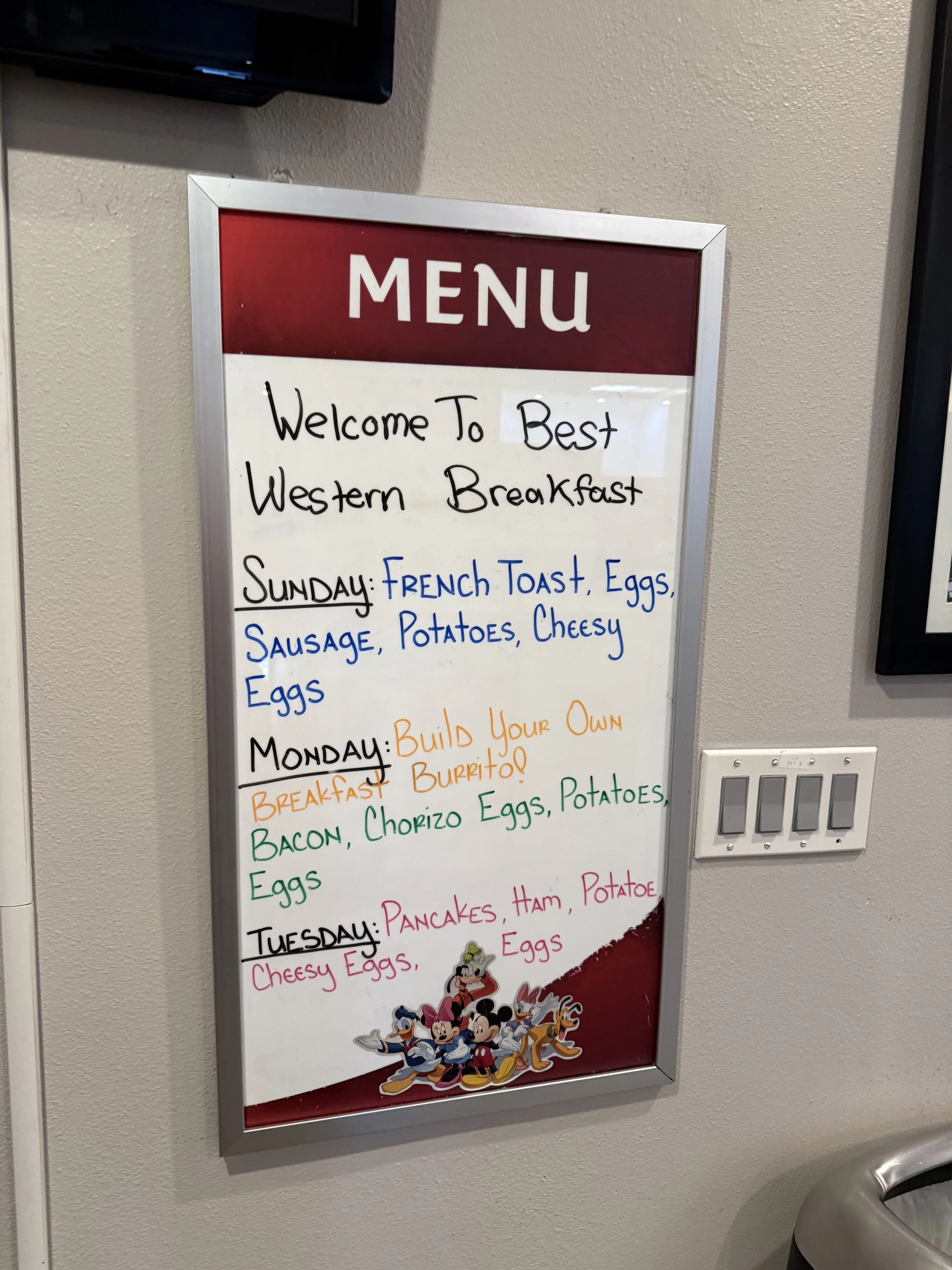 Menu board with breakfast options for Sunday, Monday, and Tuesday at a restaurant, featuring dishes like French toast, eggs, sausage, potatoes, and pancakes, with a Disney characters illustration at the bottom.