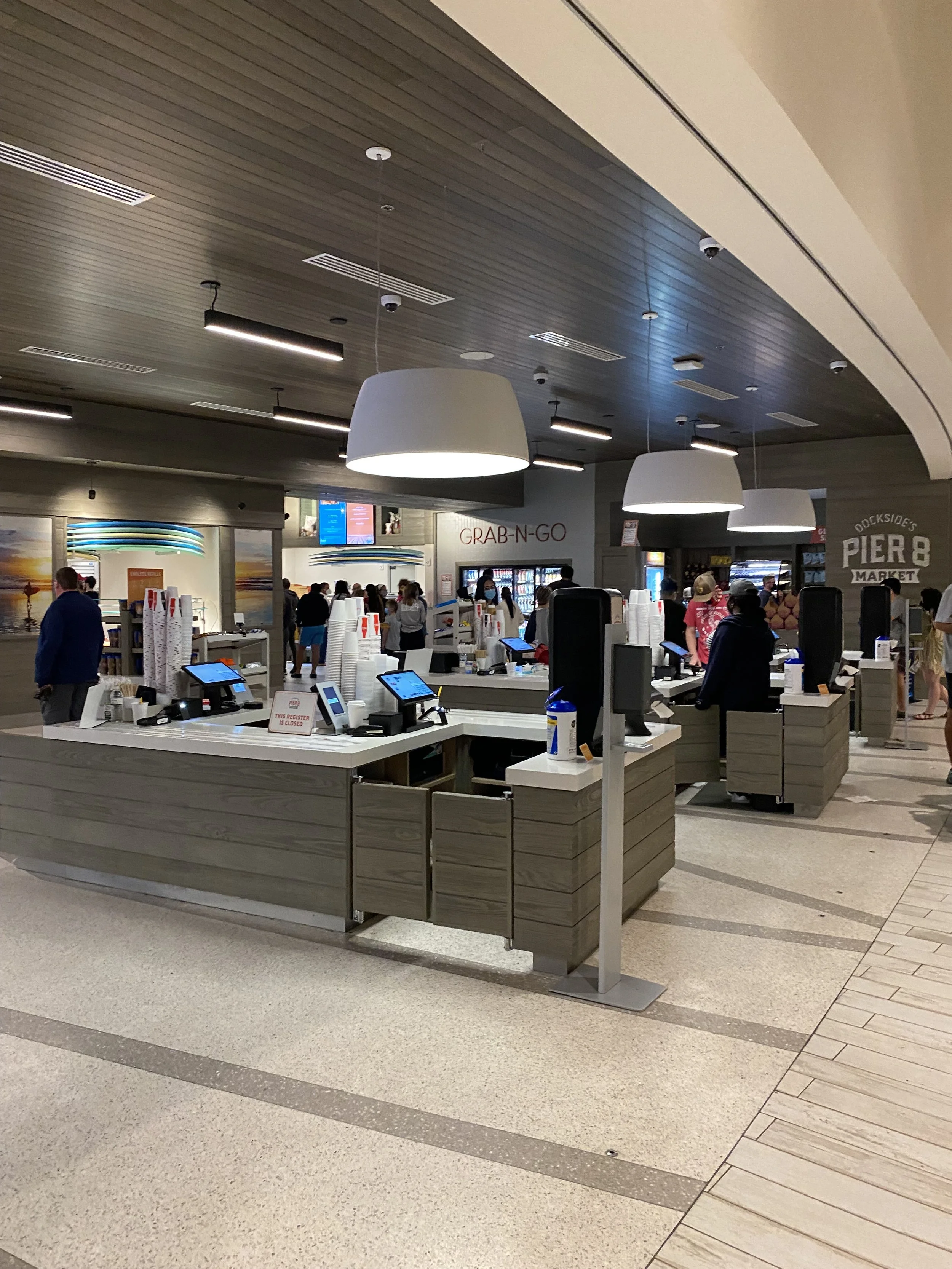 Indoor food court or market area with 'GRAB-N-GO' sign, service counters with touchscreen kiosks, and people shopping or waiting in line.