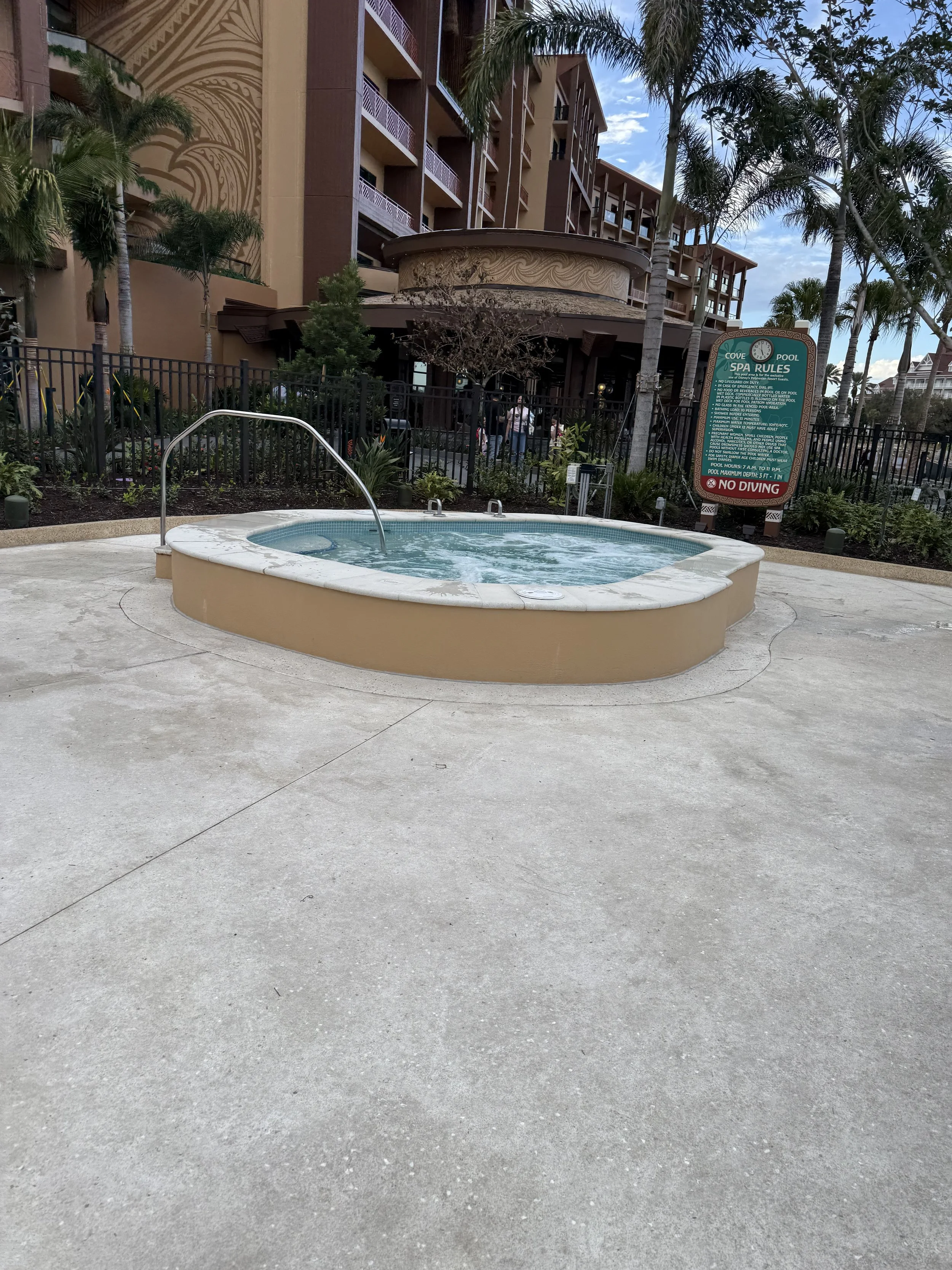 A hot tub with water, situated on a concrete pool deck, surrounded by a black fence and tall palm trees, with a building and a sign with pool rules in the background.