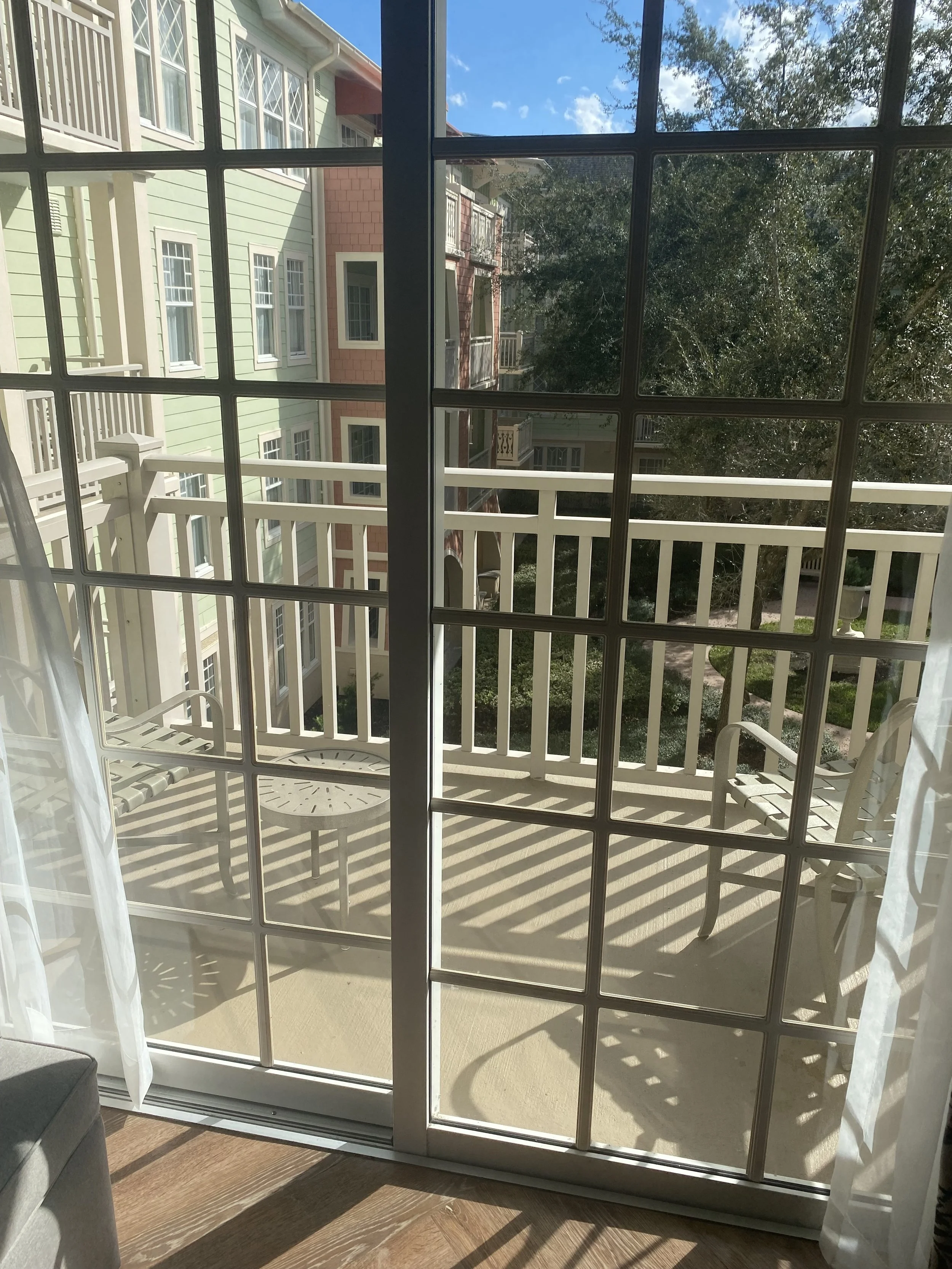 View of a balcony seen through glass sliding doors, with two white chairs and a small table, overlooking neighboring colorful residential buildings and trees under a partly cloudy sky.