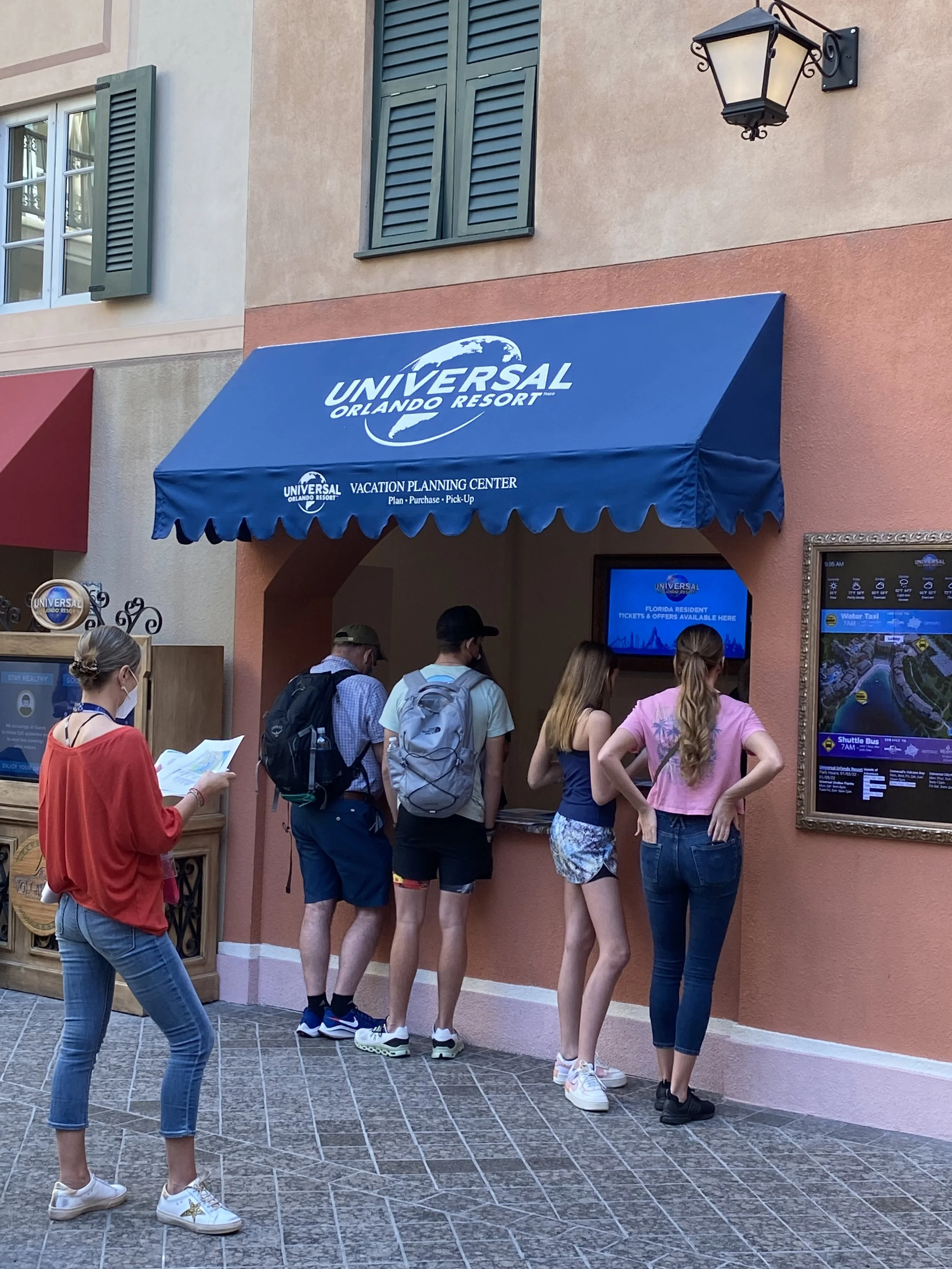 People standing in line at the ticket booth for Universal Orlando Resort, with a woman reading a brochure and others purchasing tickets, outside a colorful building with blue awning and digital displays.