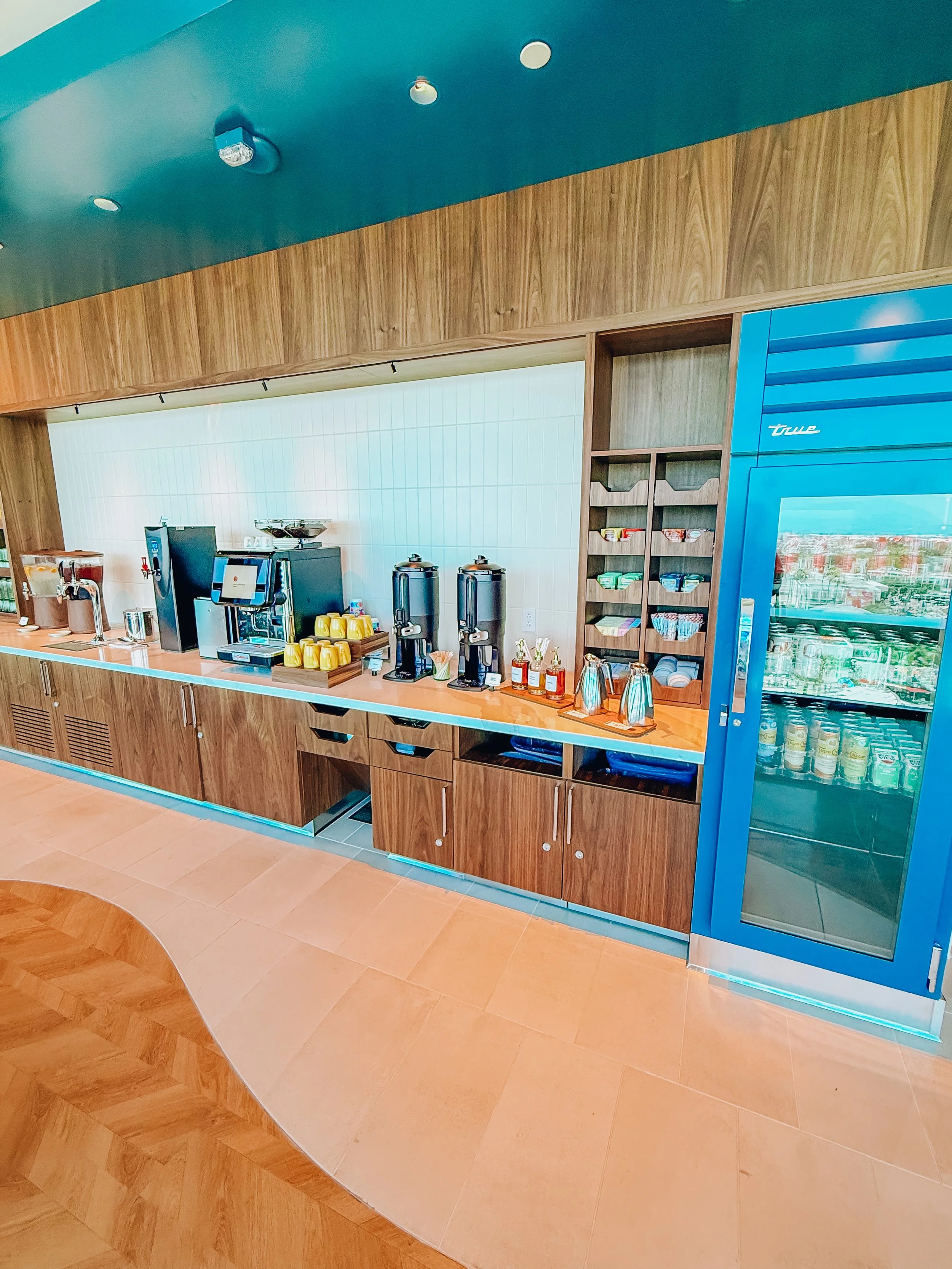 Coffee station with a coffee machine, dispensers, cups, and snacks on a wooden counter in a room with wood-paneled walls and a glass-front refrigerator.