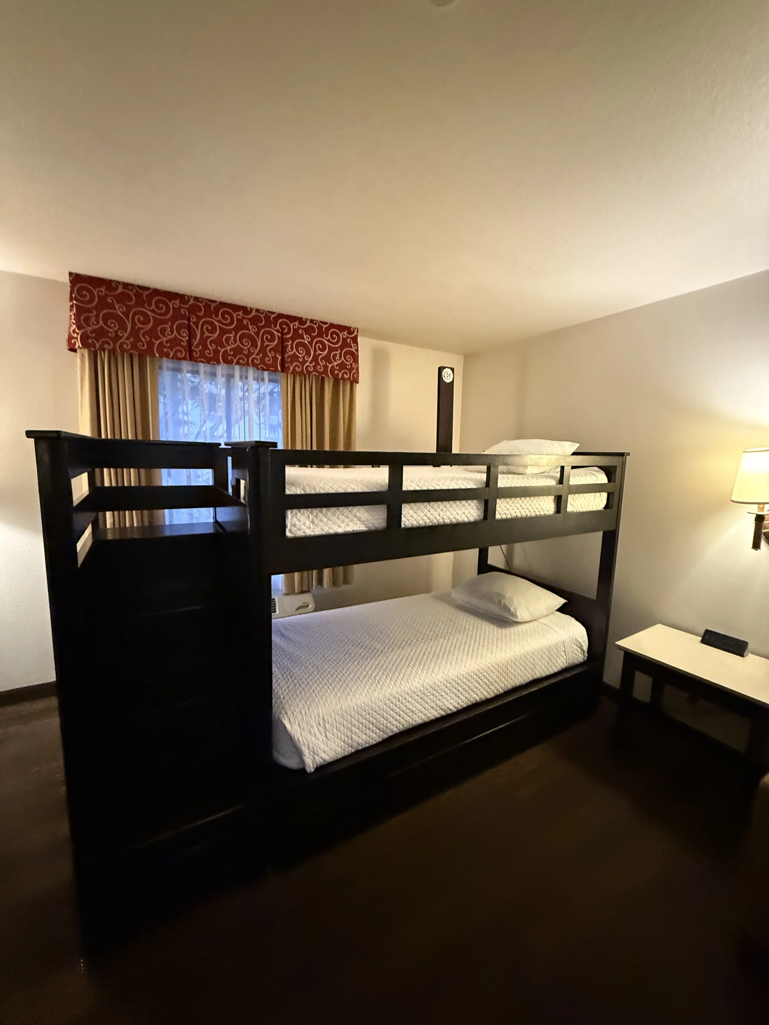 A hotel room with bunk beds, a window with curtains, a side table, and a wall lamp.