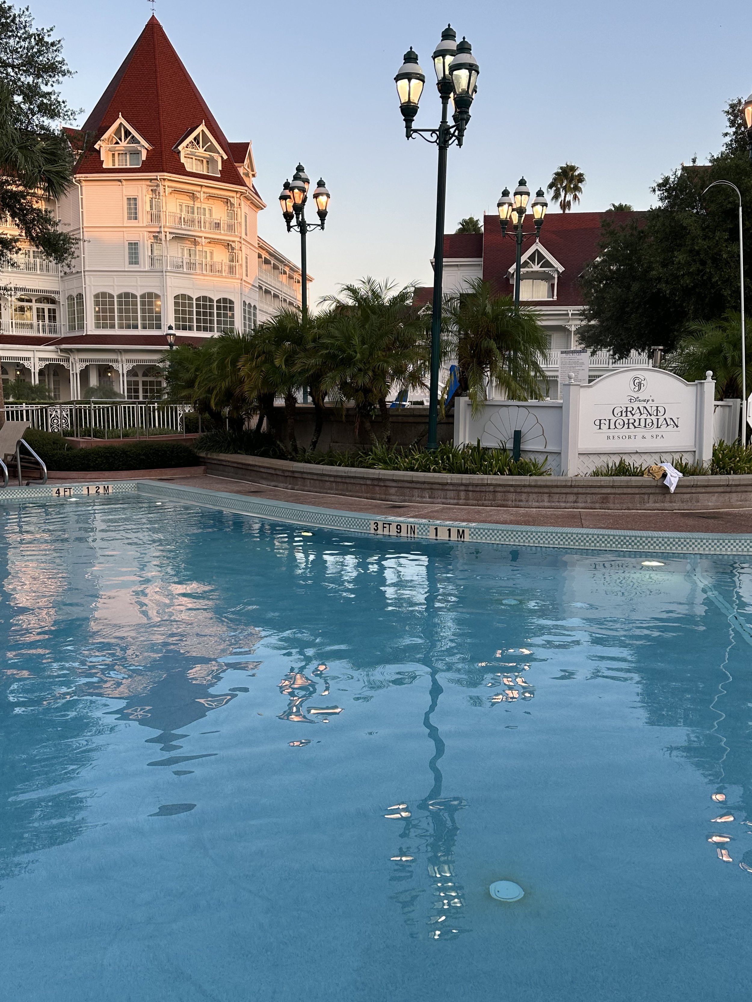 A swimming pool in front of a Victorian-style hotel with a sign that reads 'Disney's Grand Floridian Resort & Spa'. The hotel features red roofs, white siding, palm trees, and Victorian-style architecture. There are several decorative lamp posts arou