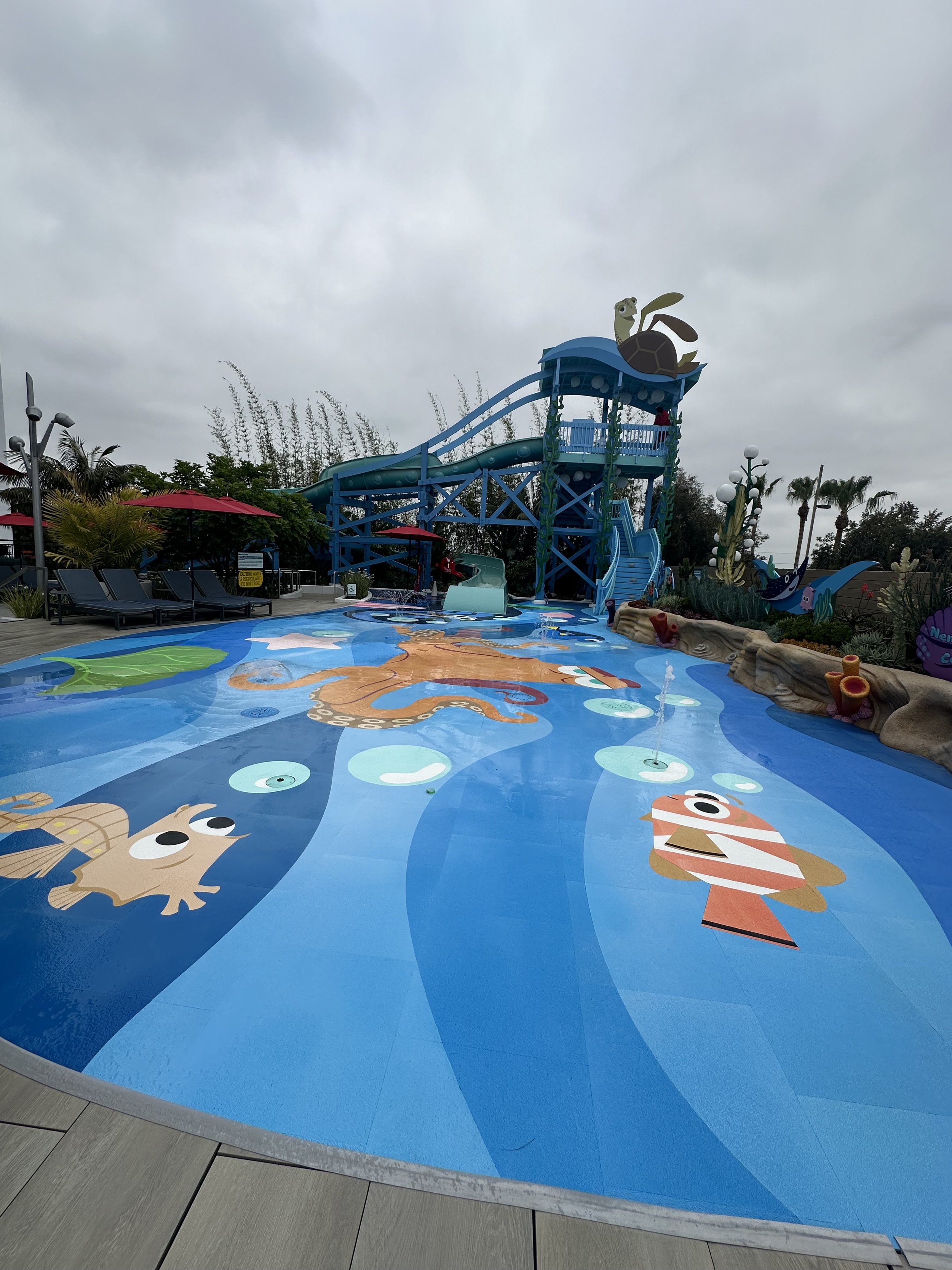 Empty splash pad with cartoon sea creatures, water slide, and shaded seating area, overcast sky.