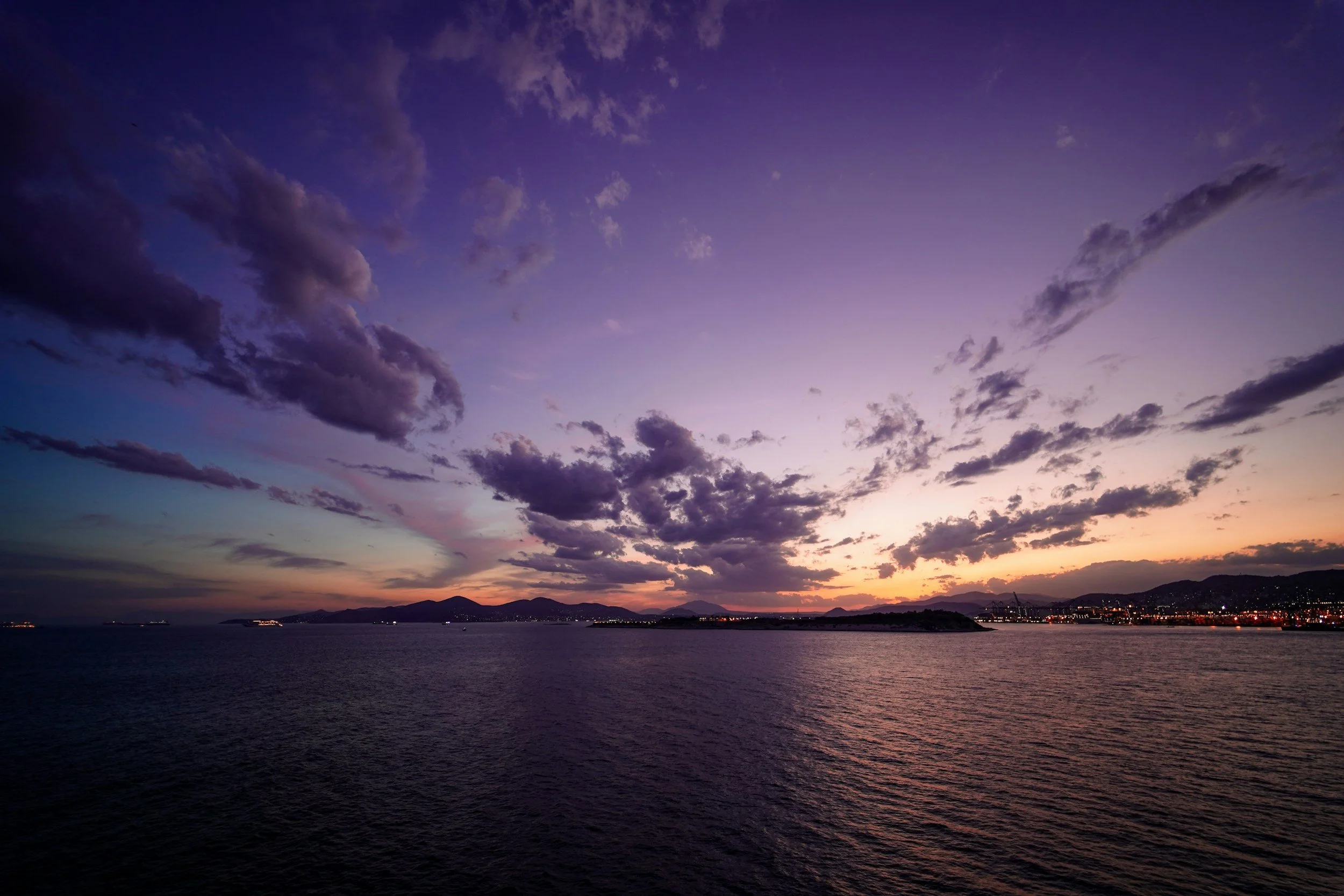A sunset over a body of water with purple, pink, and orange hues in the sky, and dark clouds scattered across the sky.