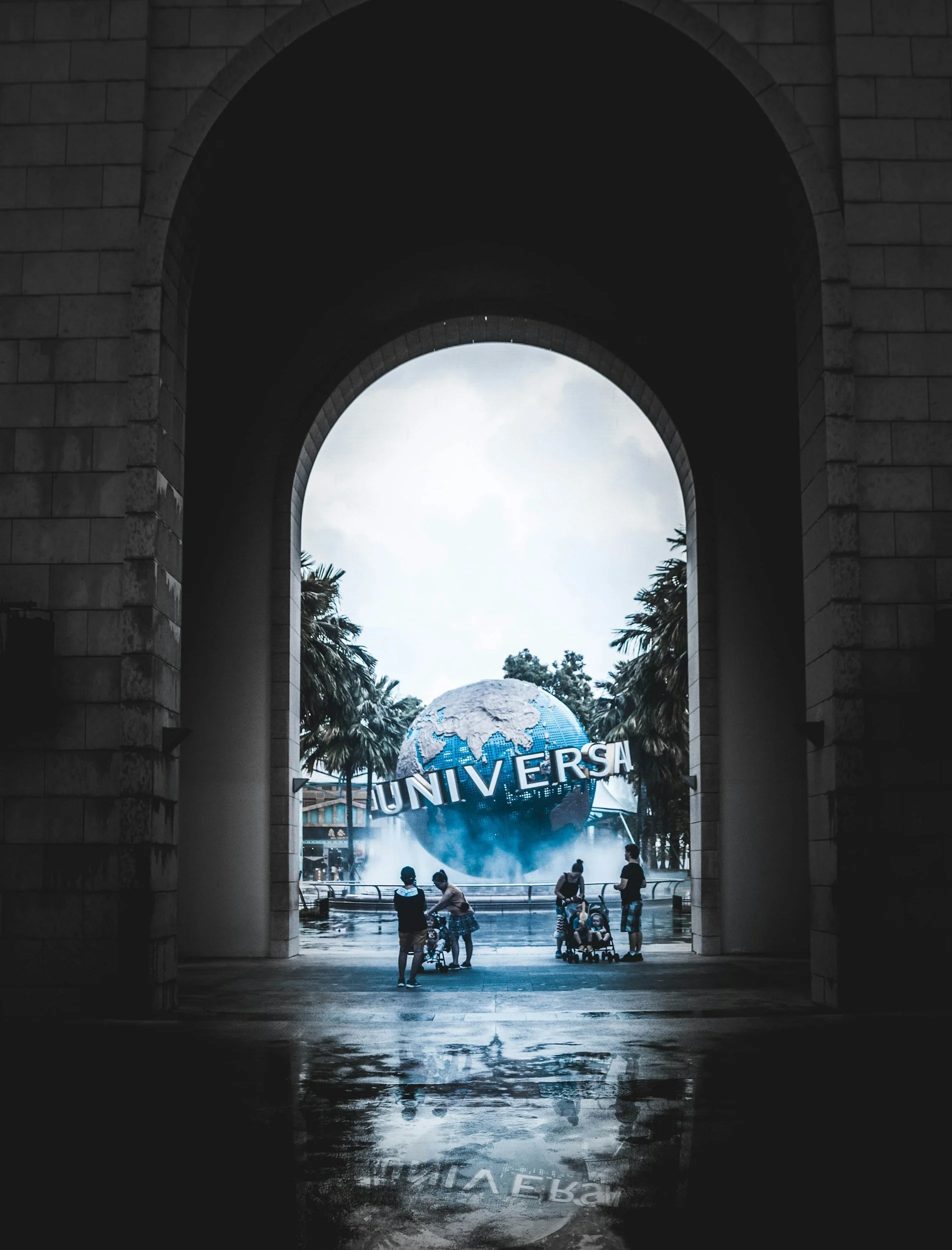 People with strollers and children standing in front of the Universal globe at a theme park, viewed through a stone archway.