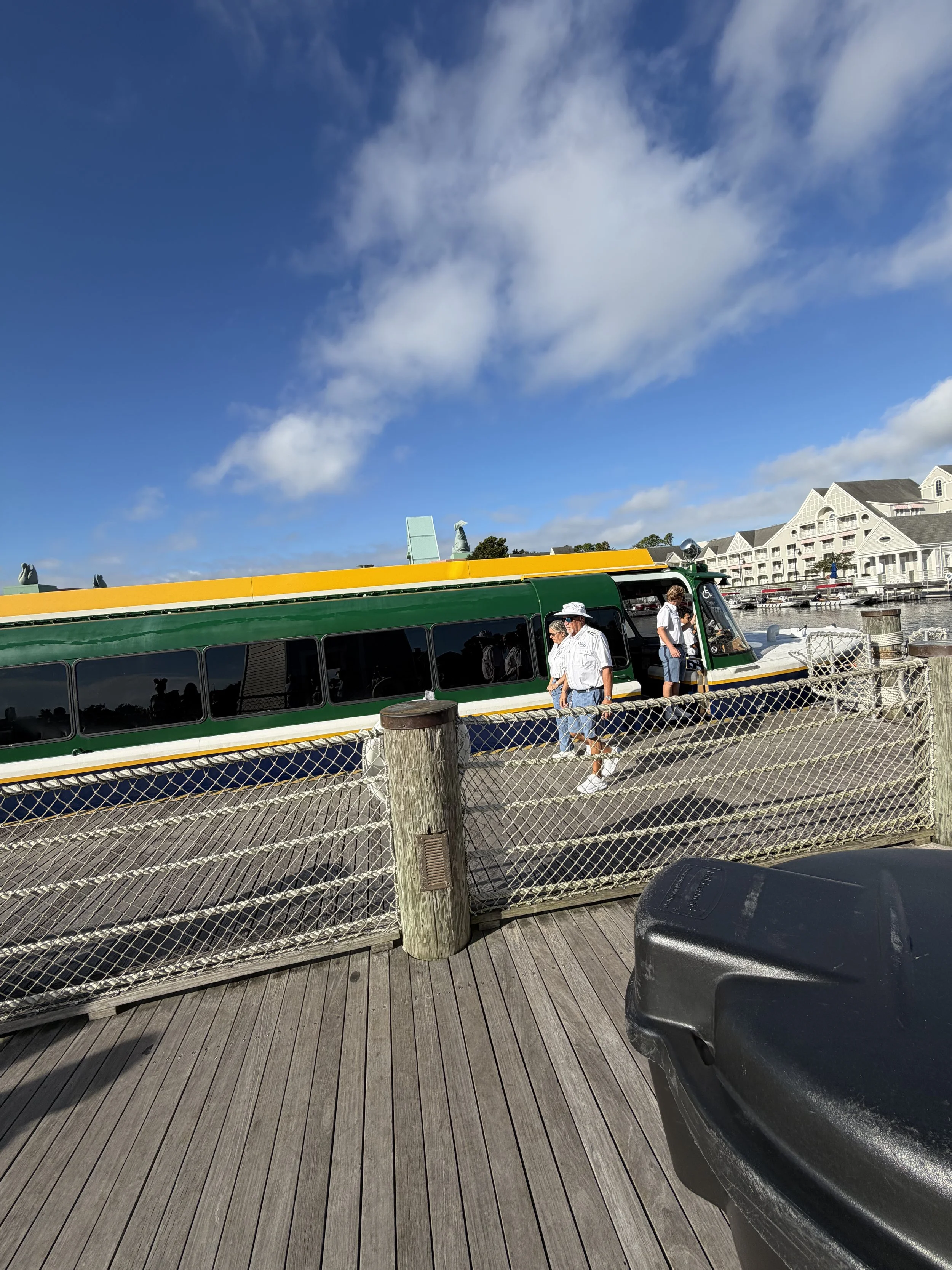 People walking on a dock beside a water taxi, with a blue sky and clouds overhead, and a large hotel building in the background.