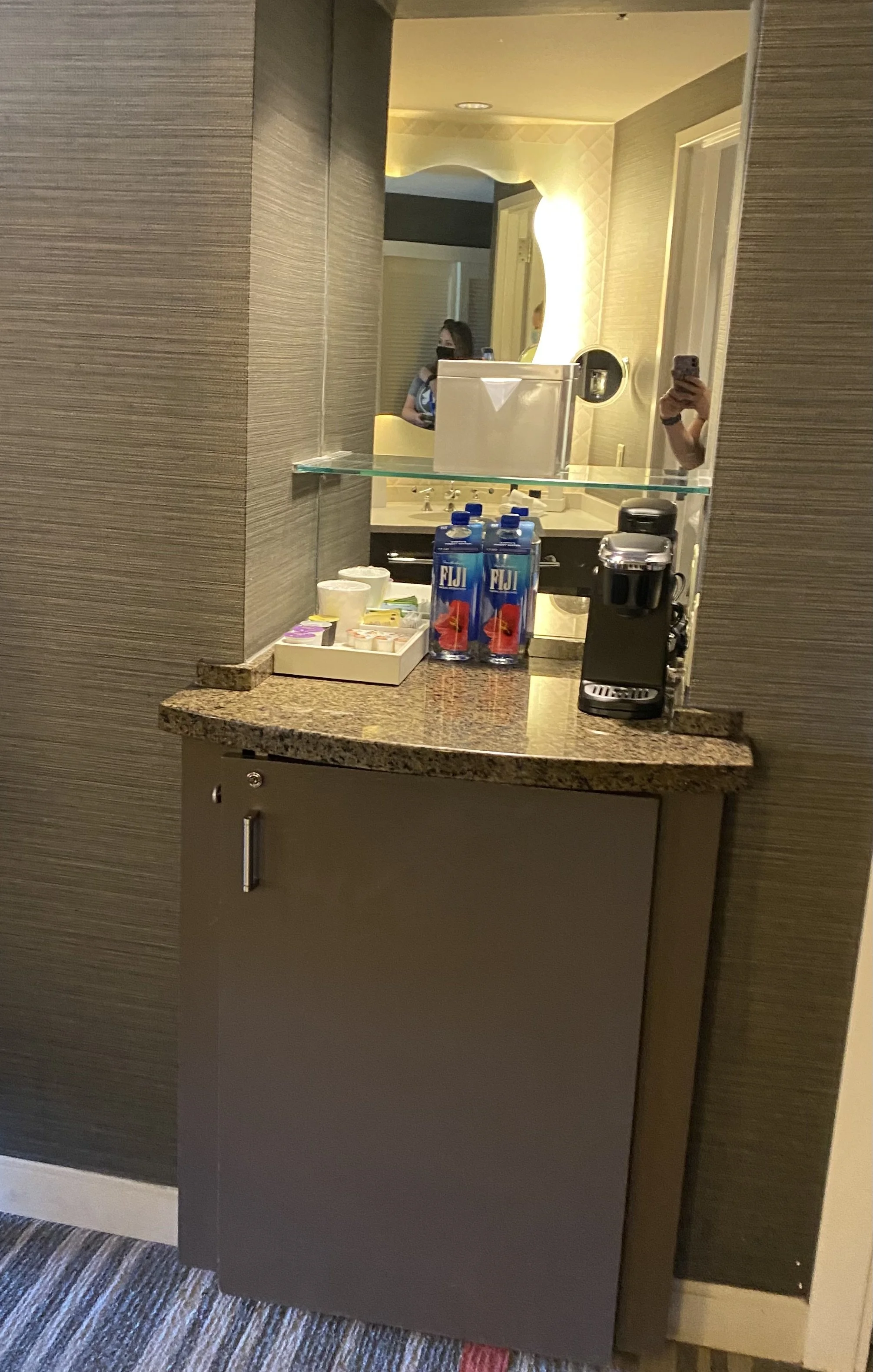 A hotel minibar area with a granite countertop, a coffee machine, three Fiji water bottles, cups, and snack packs. A mirror above the counter reflects the hotel room and a person taking a photo with a phone.