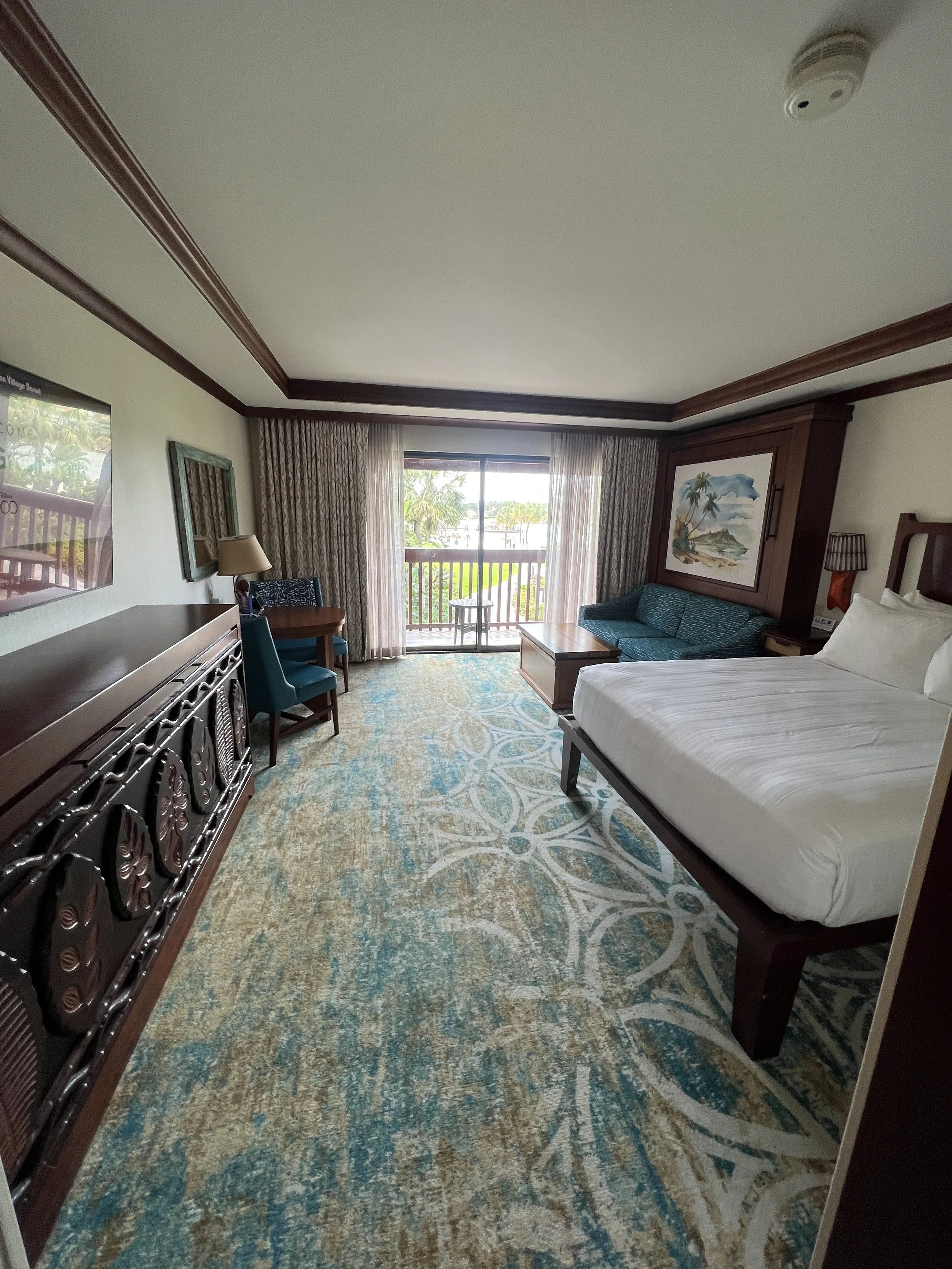 Hotel room with bed, sofa, table, chairs, and balcony view of trees and water.
