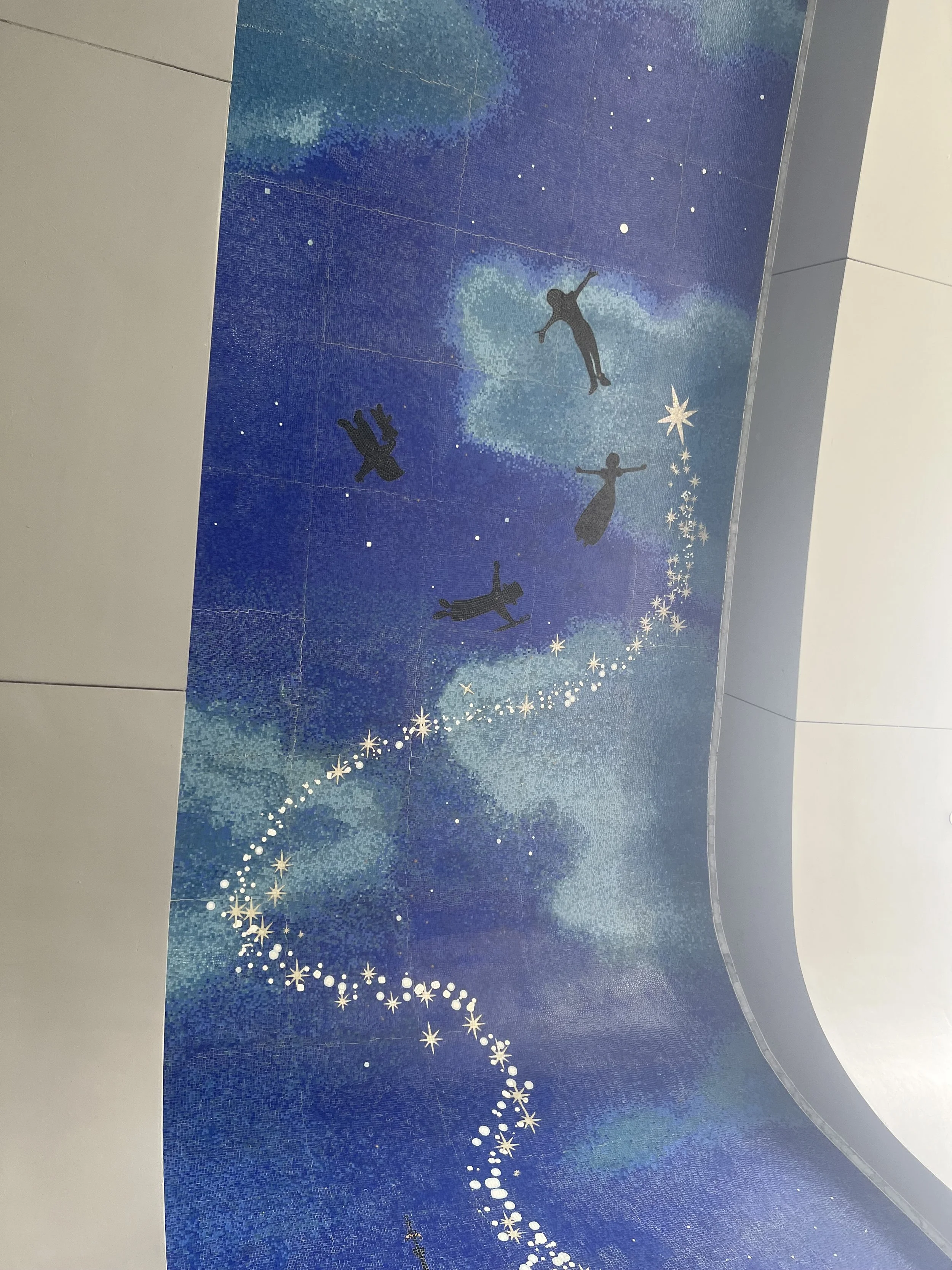 Decorative wall mural with silhouettes of four children playing, surrounded by a starry sky and swirling white star trails on a blue background.