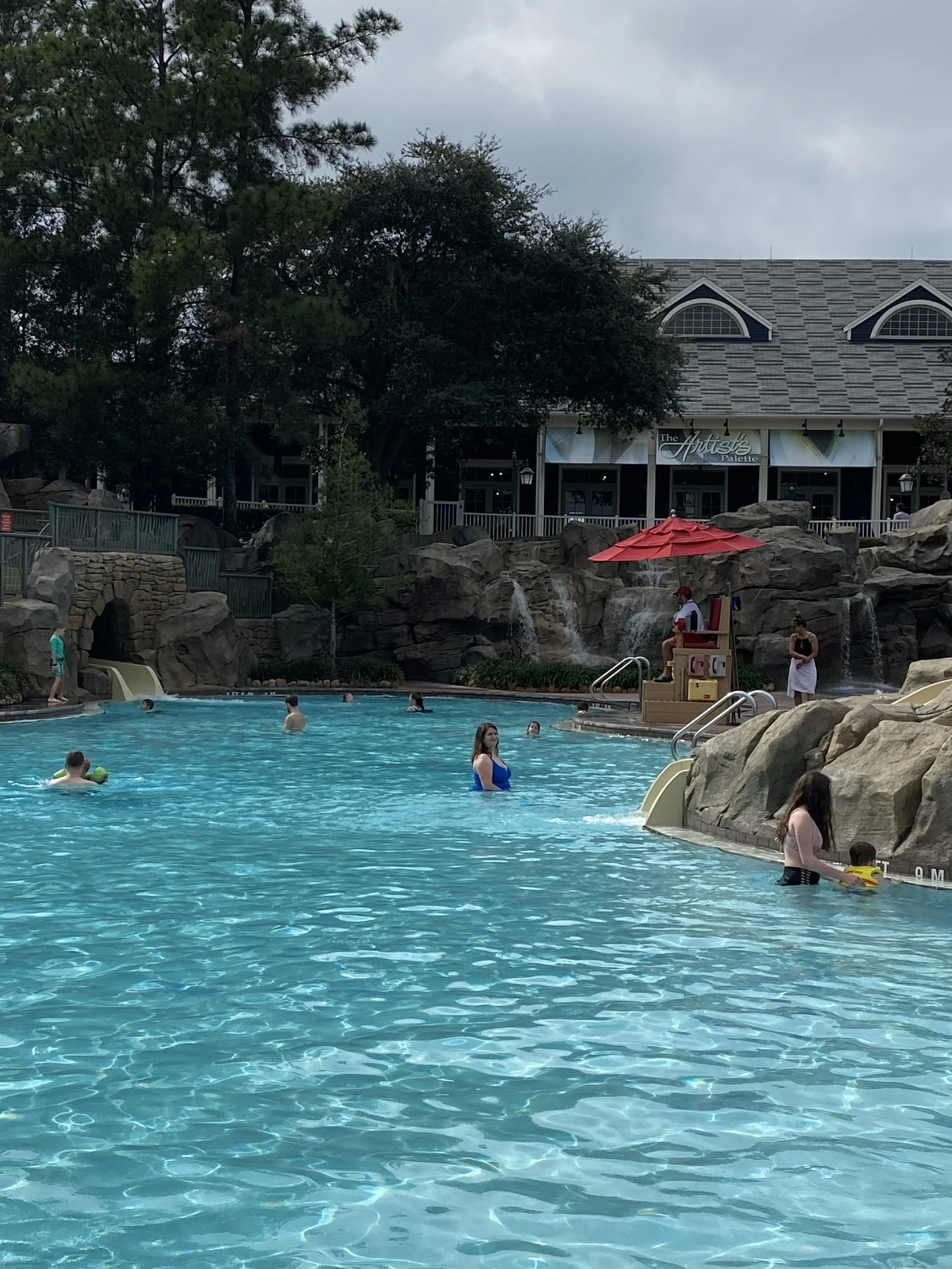 People swimming and relaxing in a large outdoor swimming pool surrounded by rocks and trees, with a building in the background.