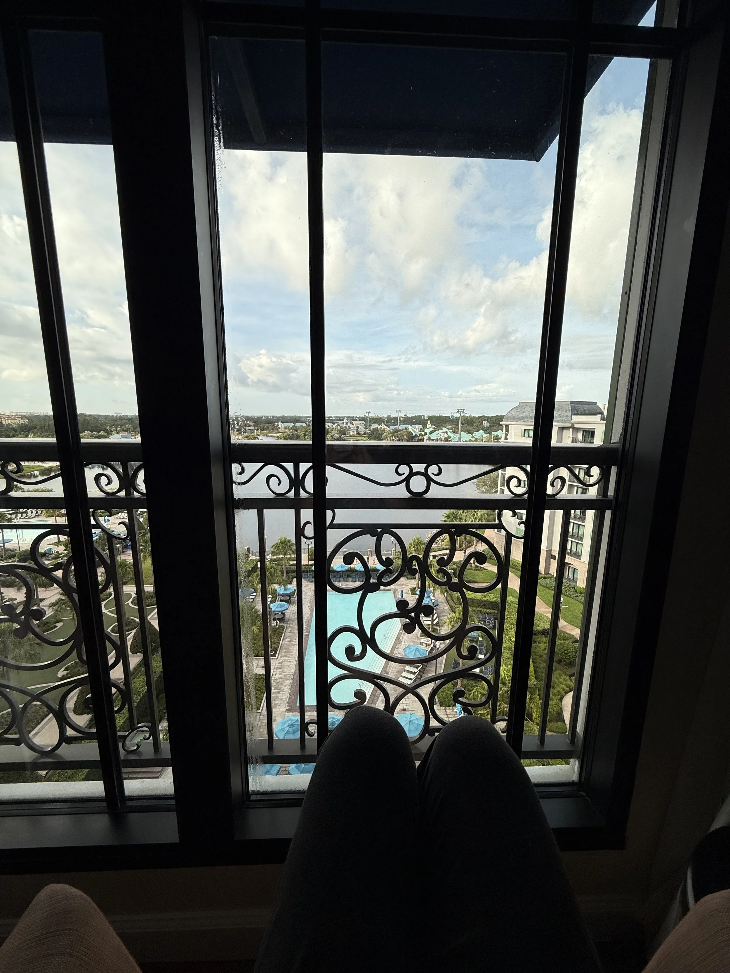 View from a high-rise balcony overlooking a swimming pool, water, buildings, and a partly cloudy sky.