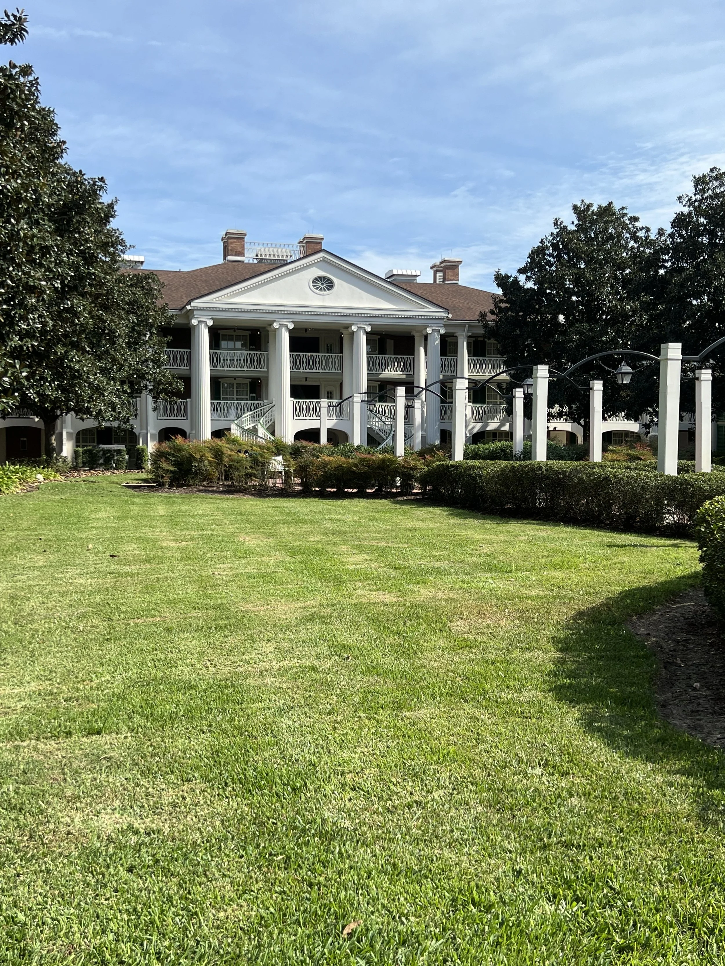 Large white mansion with columns, multiple balconies, and staircases, surrounded by a well-manicured lawn and trees.