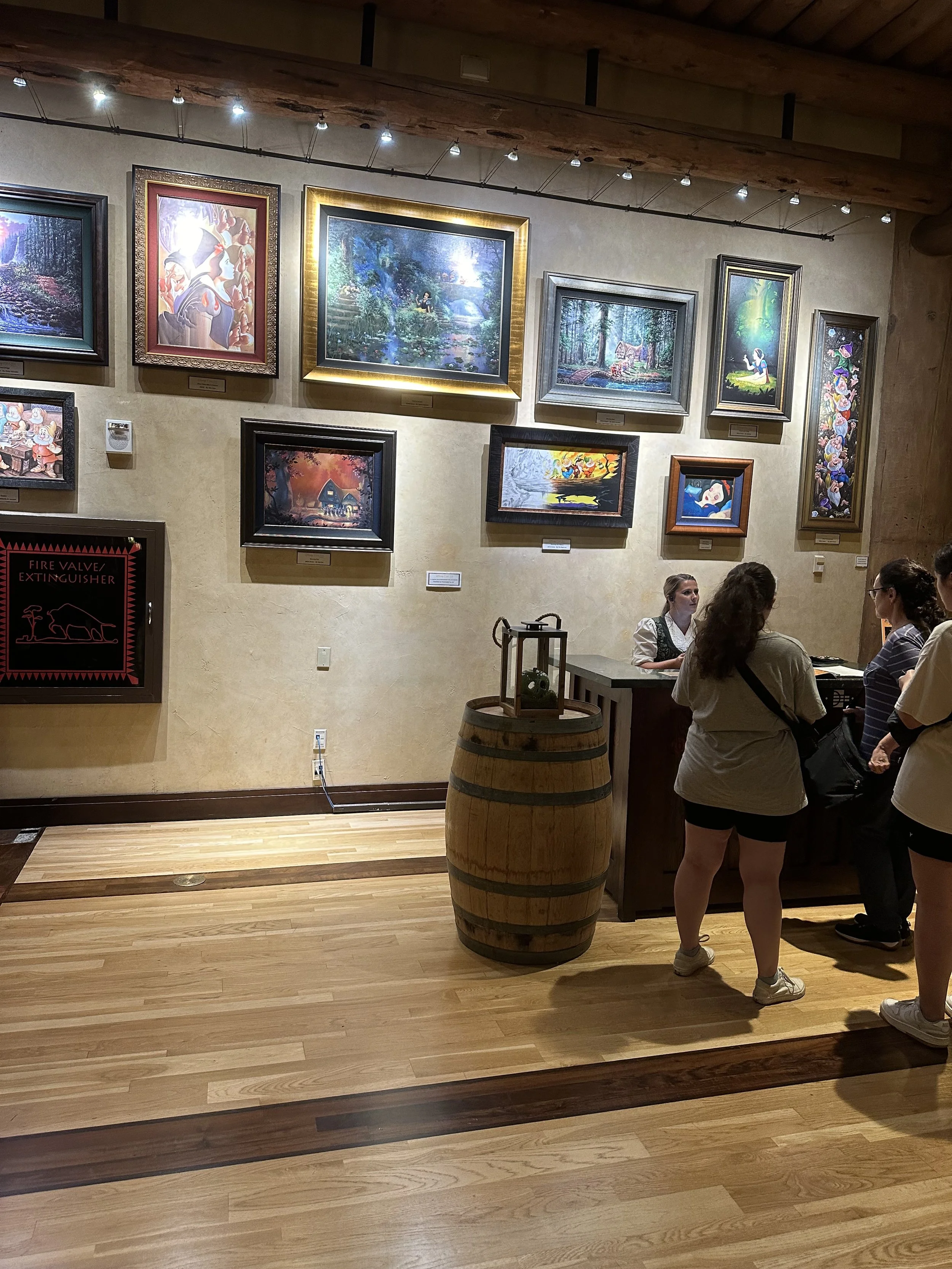 People looking at paintings in an art gallery with wooden floors and a beige wall. There is a barrel with a lantern on top near the reception desk.