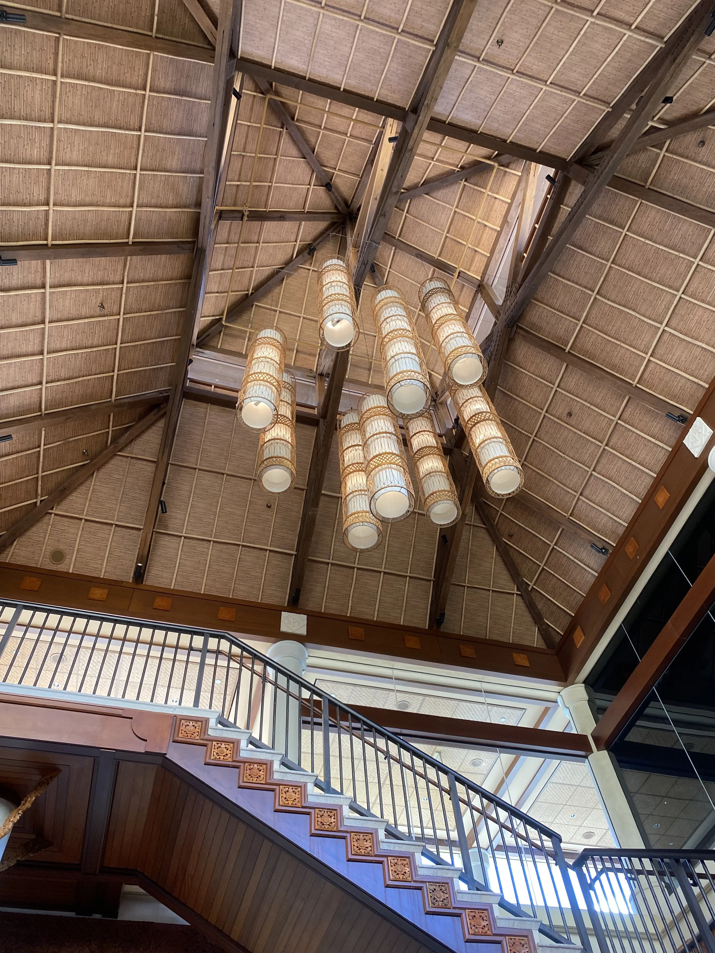 Interior view of a high ceiling with wooden beams and a cluster of hanging cylindrical lamps, with a staircase featuring wooden railings and decorative panels.