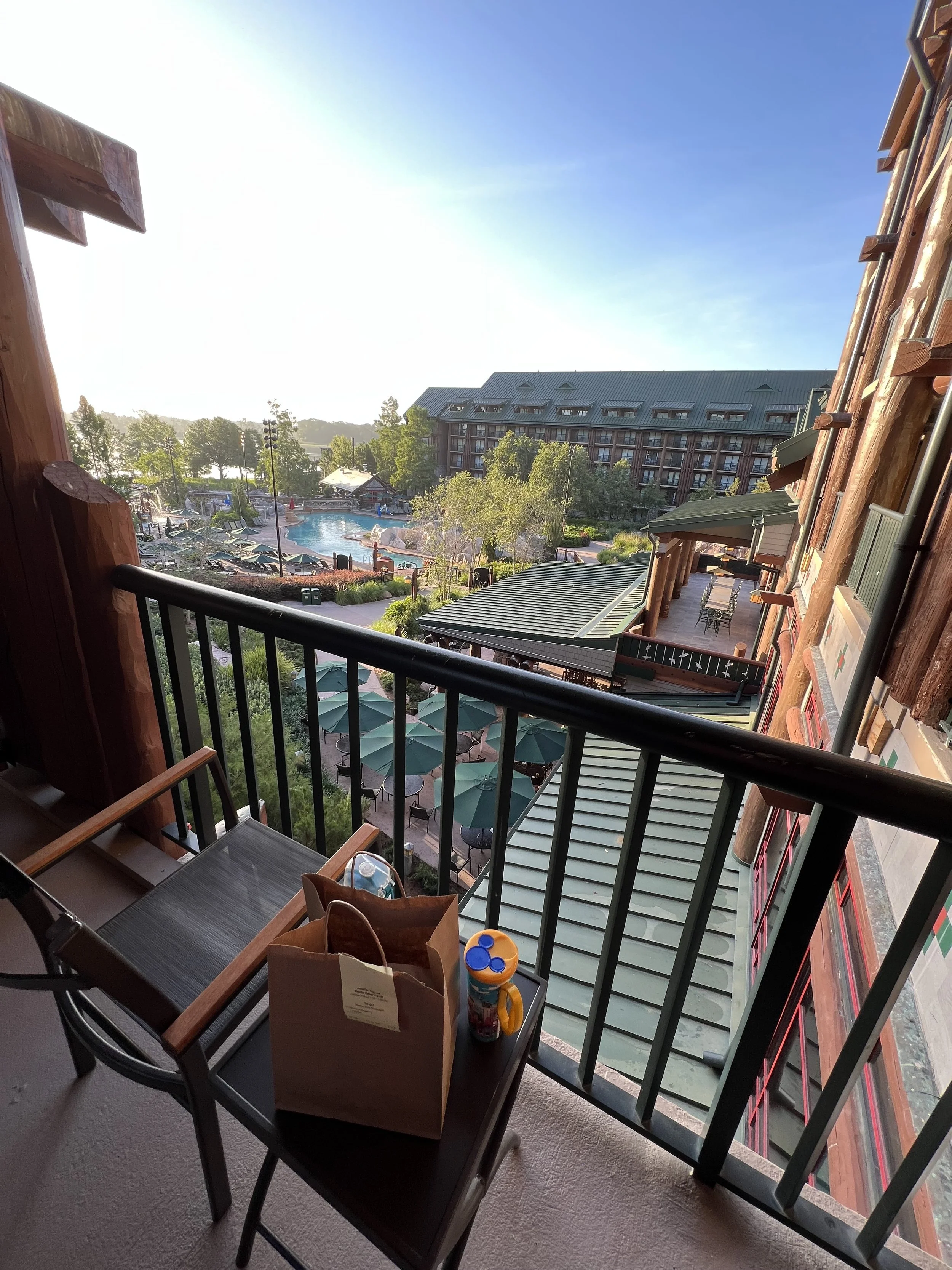View from a balcony overlooking a pool area with lounge chairs and green umbrellas, surrounded by lush trees and a large multi-story building under a clear blue sky.