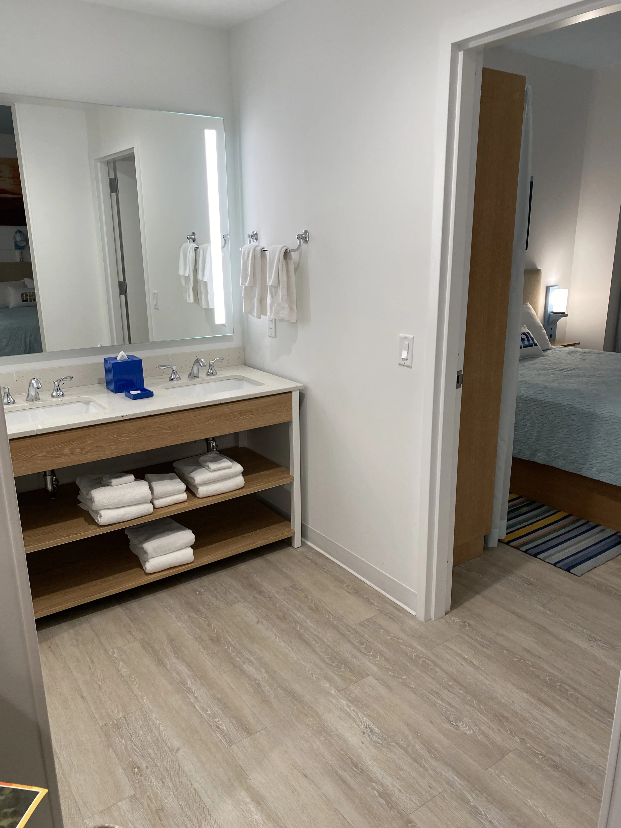 Bathroom with double sink vanity, large mirror, towels on towel racks, and a view into a bedroom with a bed, nightstand, and lamp.