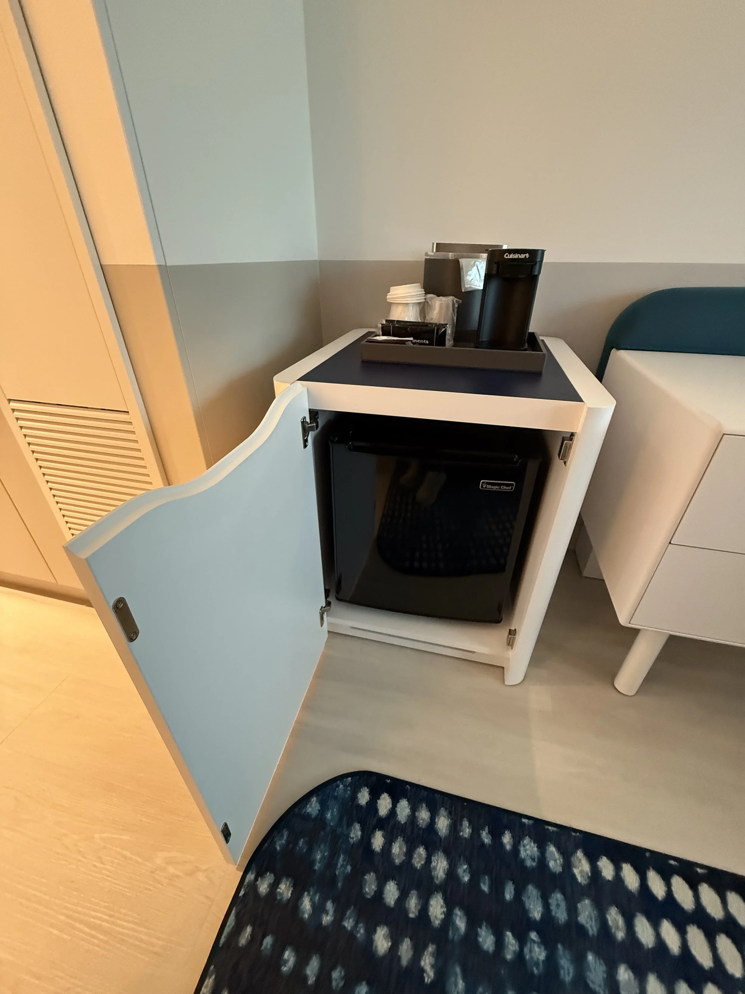 Mini refrigerator with open door in a hotel room, next to a white cabinet and a blue rug.