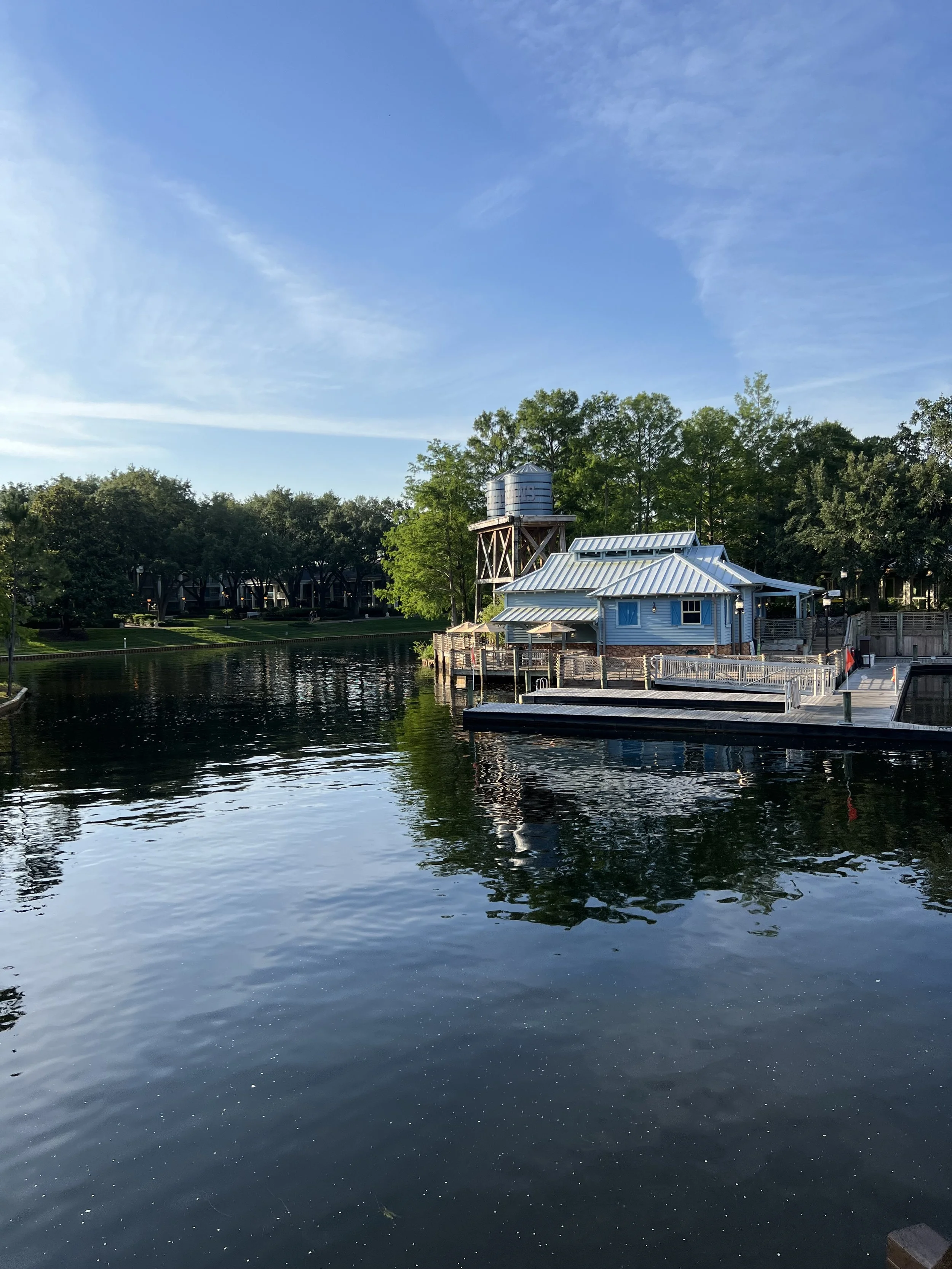 A peaceful waterway with a docked blue building, water tower, and lush green trees in the background, under a clear blue sky.