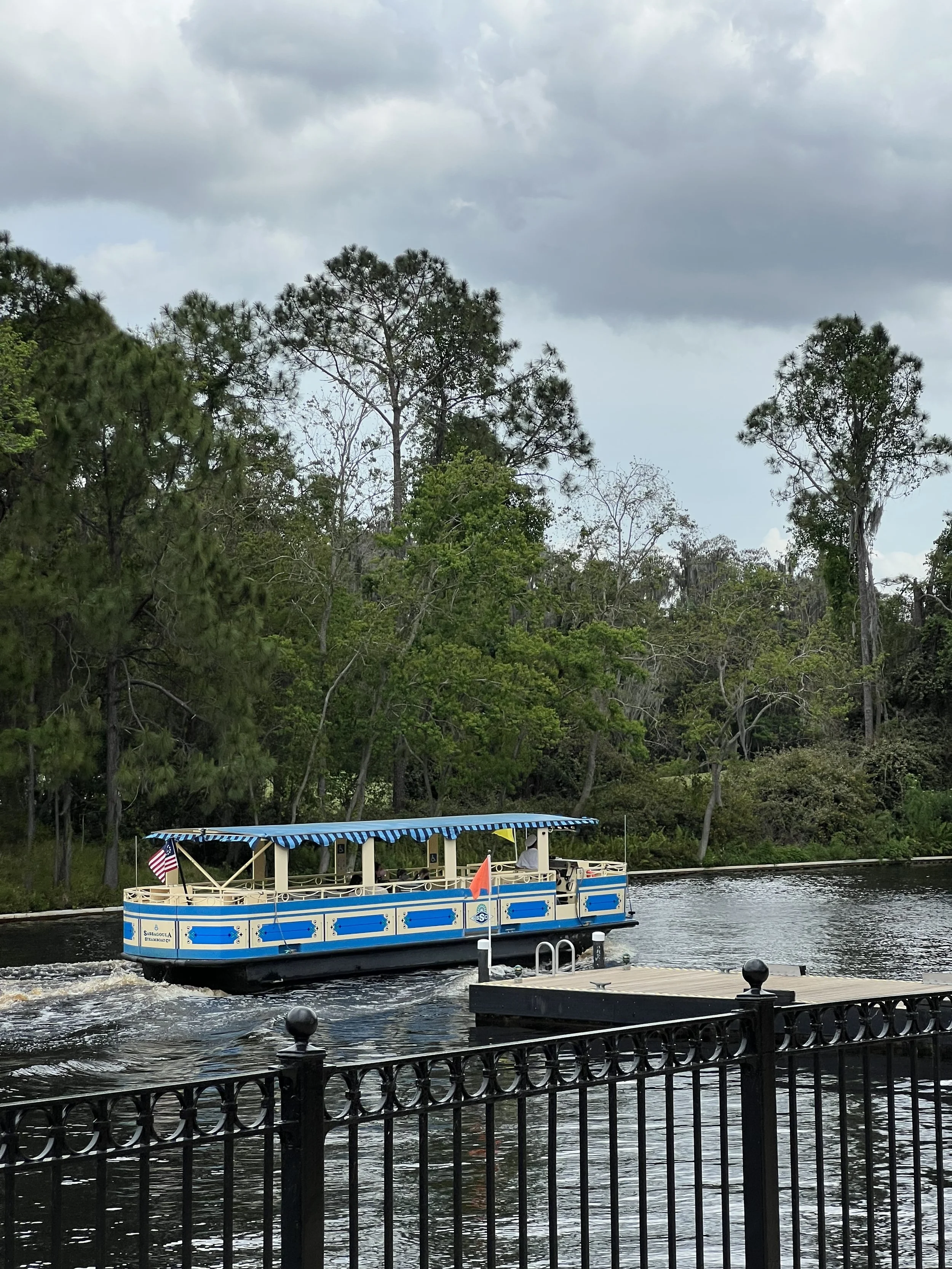 A small boat with a blue and white striped canopy floating on a river, with trees in the background and a cloudy sky overhead.