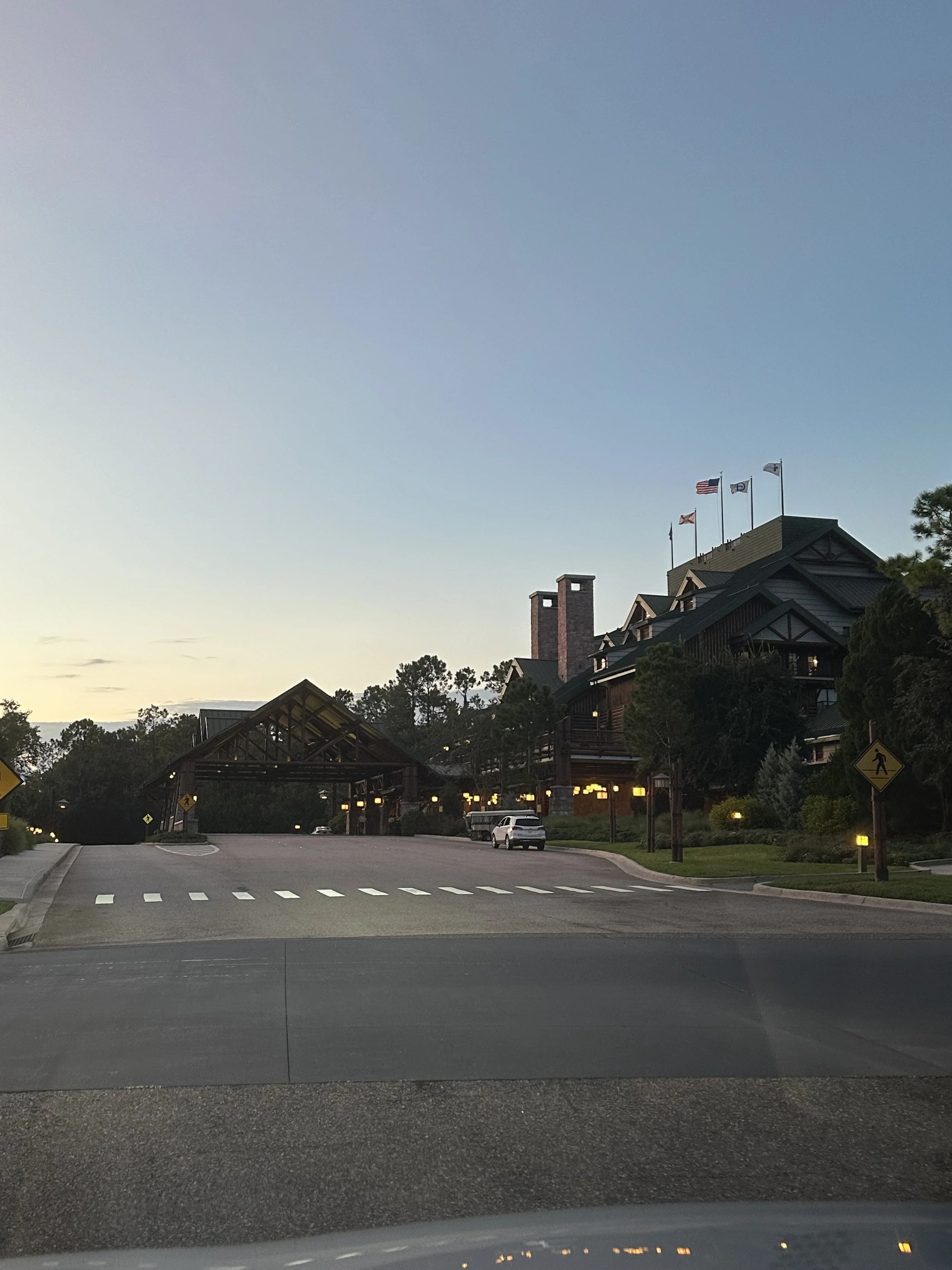 Entrance to a lodge or hotel with a covered driveway, surrounded by trees and greenery, with flags flying on the roof, and street signs and parked cars visible.